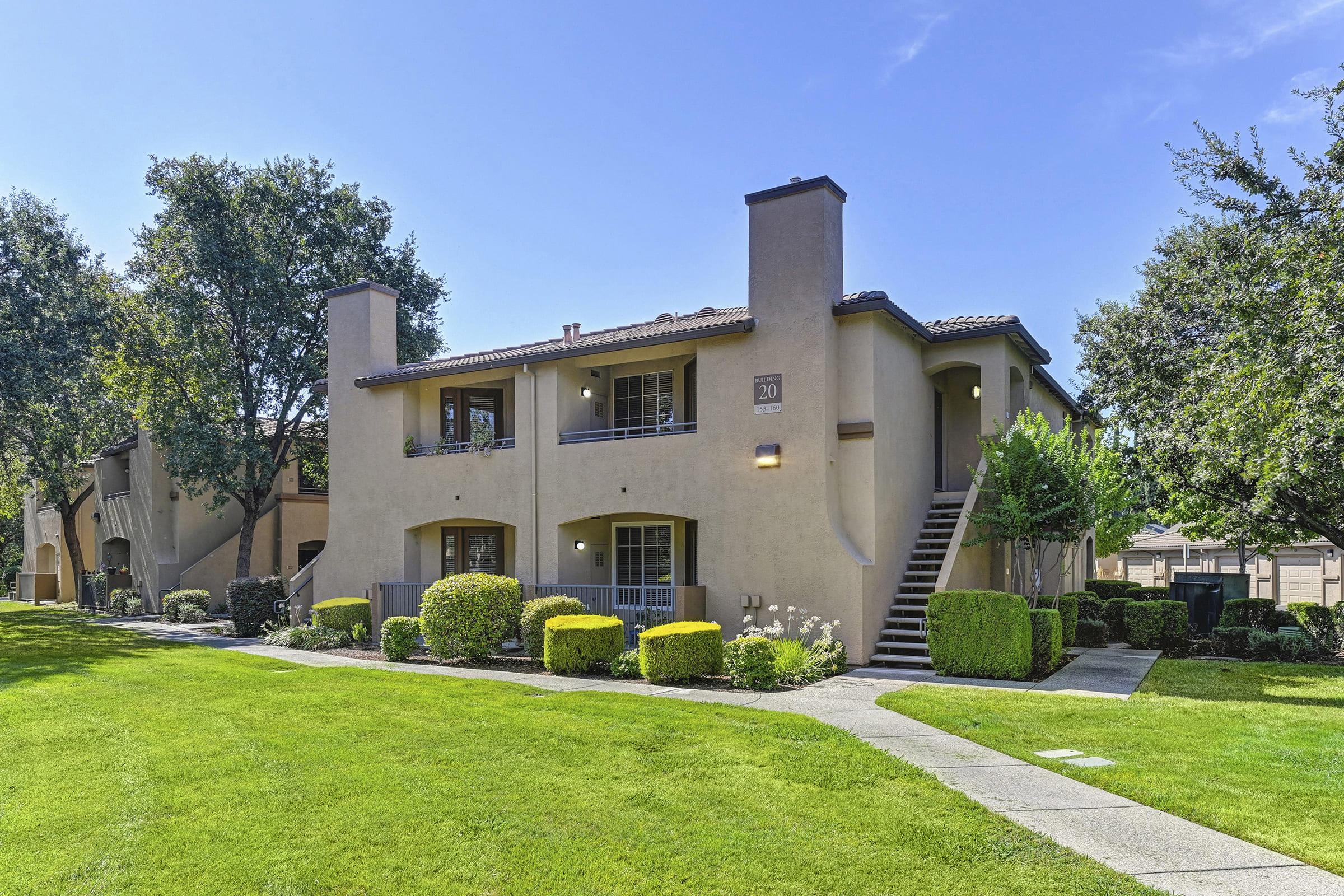 Two-story apartment building with a beige exterior, featuring balconies and green landscaping. A walkway leads to the entrance, surrounded by well-maintained lawns and shrubs. Trees provide shade in the area, under a clear blue sky.
