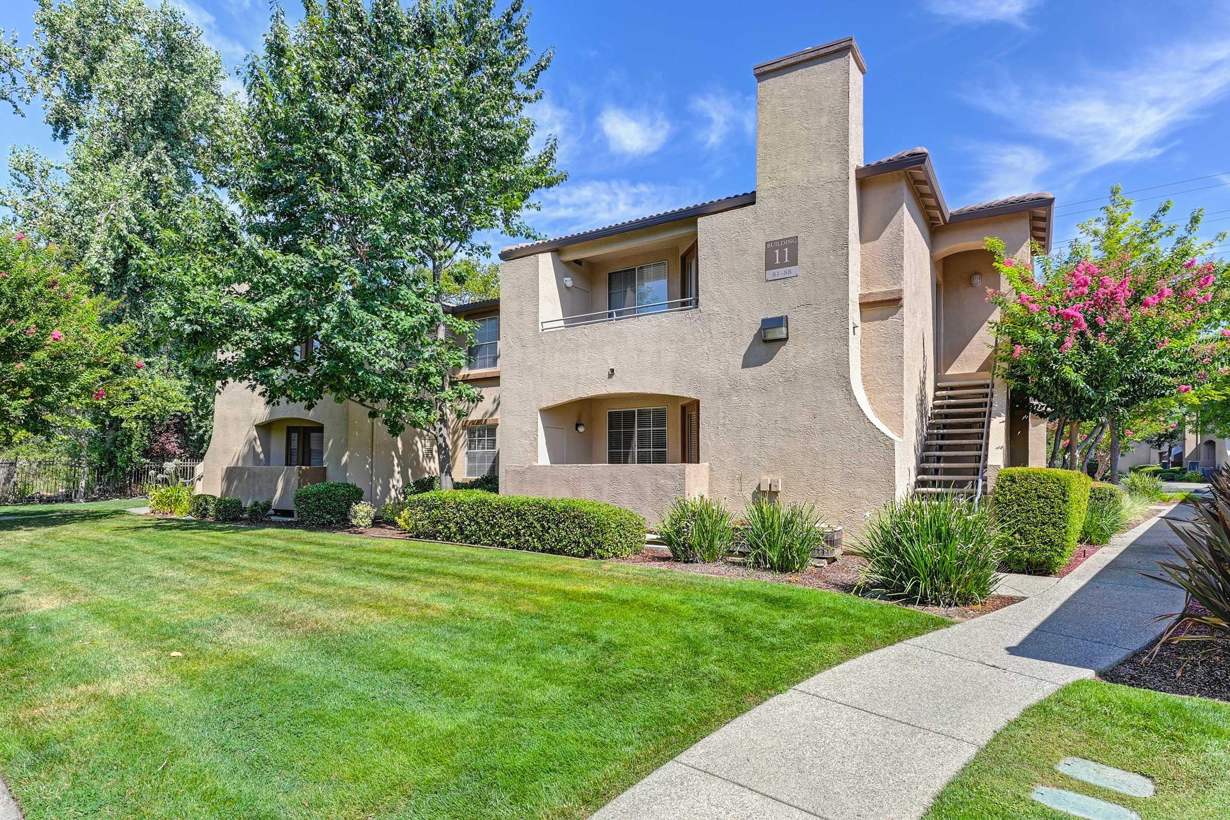 Two-story apartment building surrounded by well-maintained lawns and landscaping. A pathway leads to the entrance, and there are trees and flowering plants nearby. The sky is clear with a few clouds, indicating a sunny day.