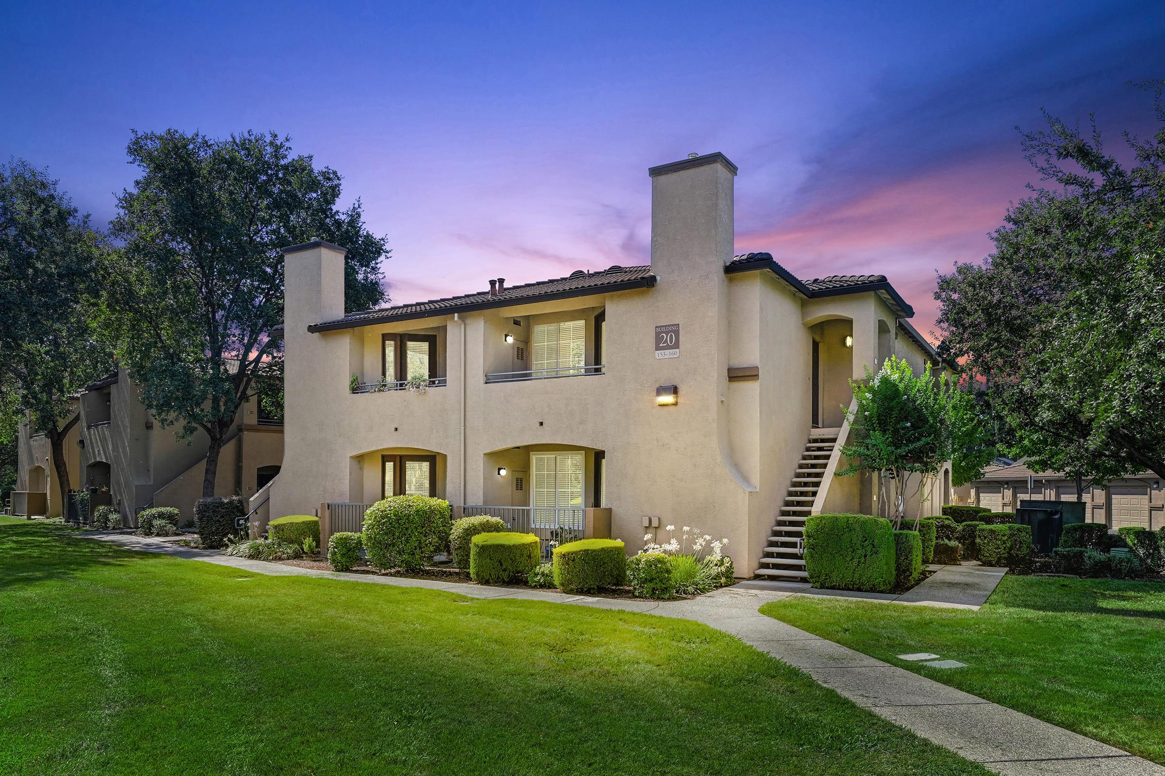 Two-story apartment building surrounded by well-maintained green lawns and shrubs. The building features a light-colored exterior with balconies and a staircase leading to the second floor. A pastel sunset sky with purple and orange hues is visible in the background.