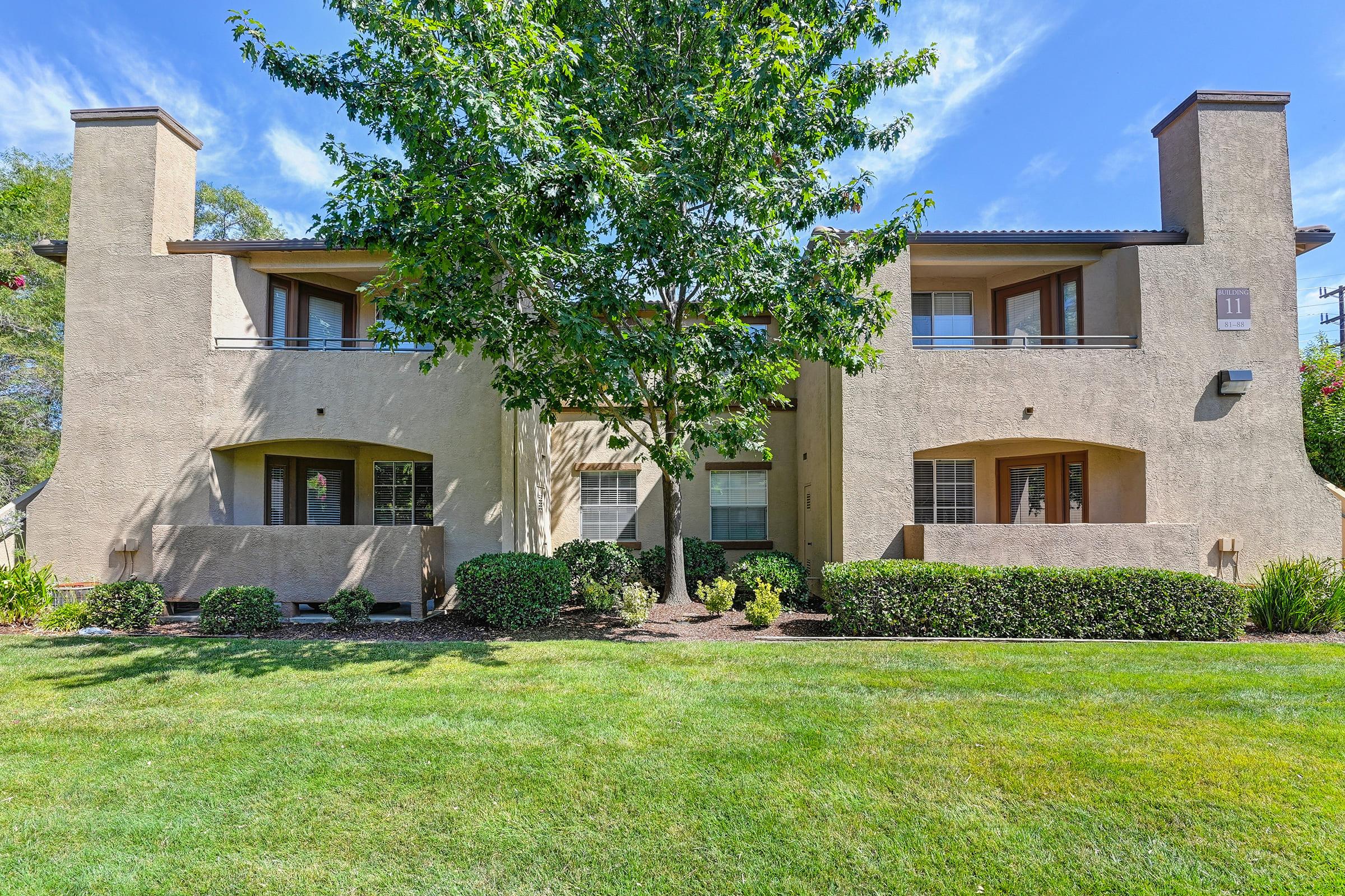 A two-story building with a beige exterior, featuring two separate units. Each unit has large windows and a patio area. The front yard is well-maintained with grass and shrubs, and a large tree sits in the center. The sky is clear with a few clouds.