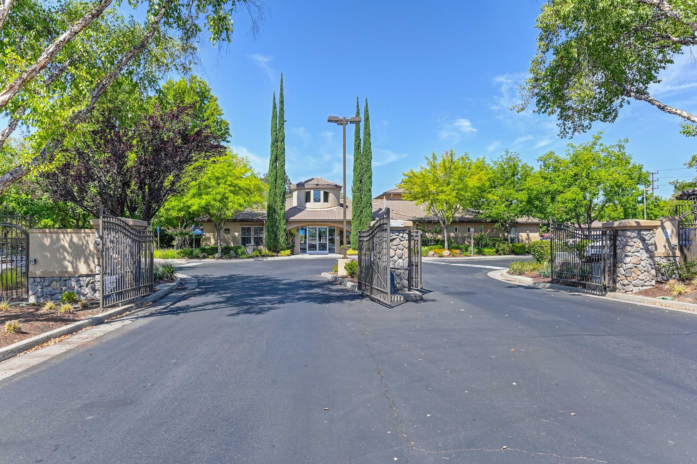 Entrance to a gated community featuring a curved road leading to a clubhouse. Flanked by tall trees and landscaped greenery, the scene is set under a clear blue sky. The gates are partially open, inviting residents or guests into the area.