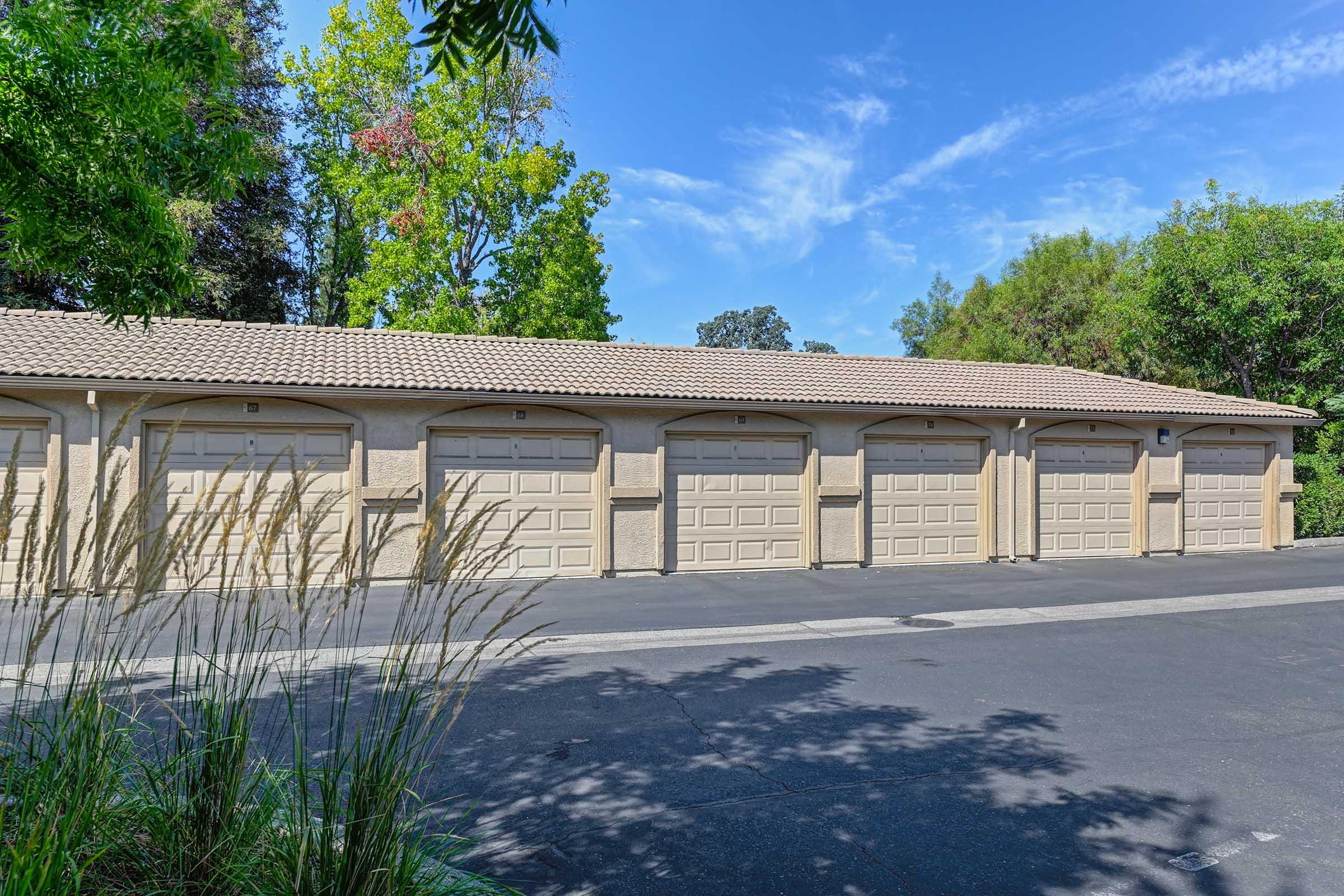 A row of beige garage doors with a tiled roof, surrounded by green trees and shrubs under a clear blue sky. The driveway is paved, and ornamental grass is visible in the foreground, adding a touch of nature to the scene.