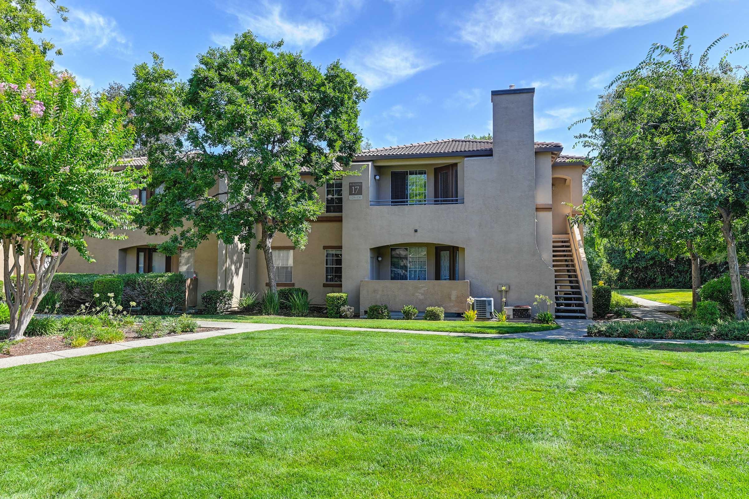 A well-maintained residential building surrounded by green lawns and trees. The property features a two-story structure with balconies, a walkway, and landscaped areas. Bright blue sky with scattered clouds creates a pleasant atmosphere.