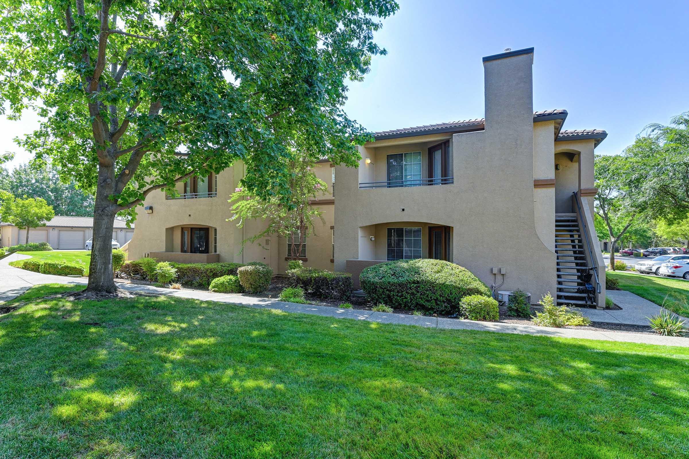 A two-story apartment building with a stucco exterior, surrounded by well-maintained greenery and trees. A pathway leads to the entrance, and the building features balconies on the upper level. There is a parking area visible in the background. The setting is sunny and inviting.
