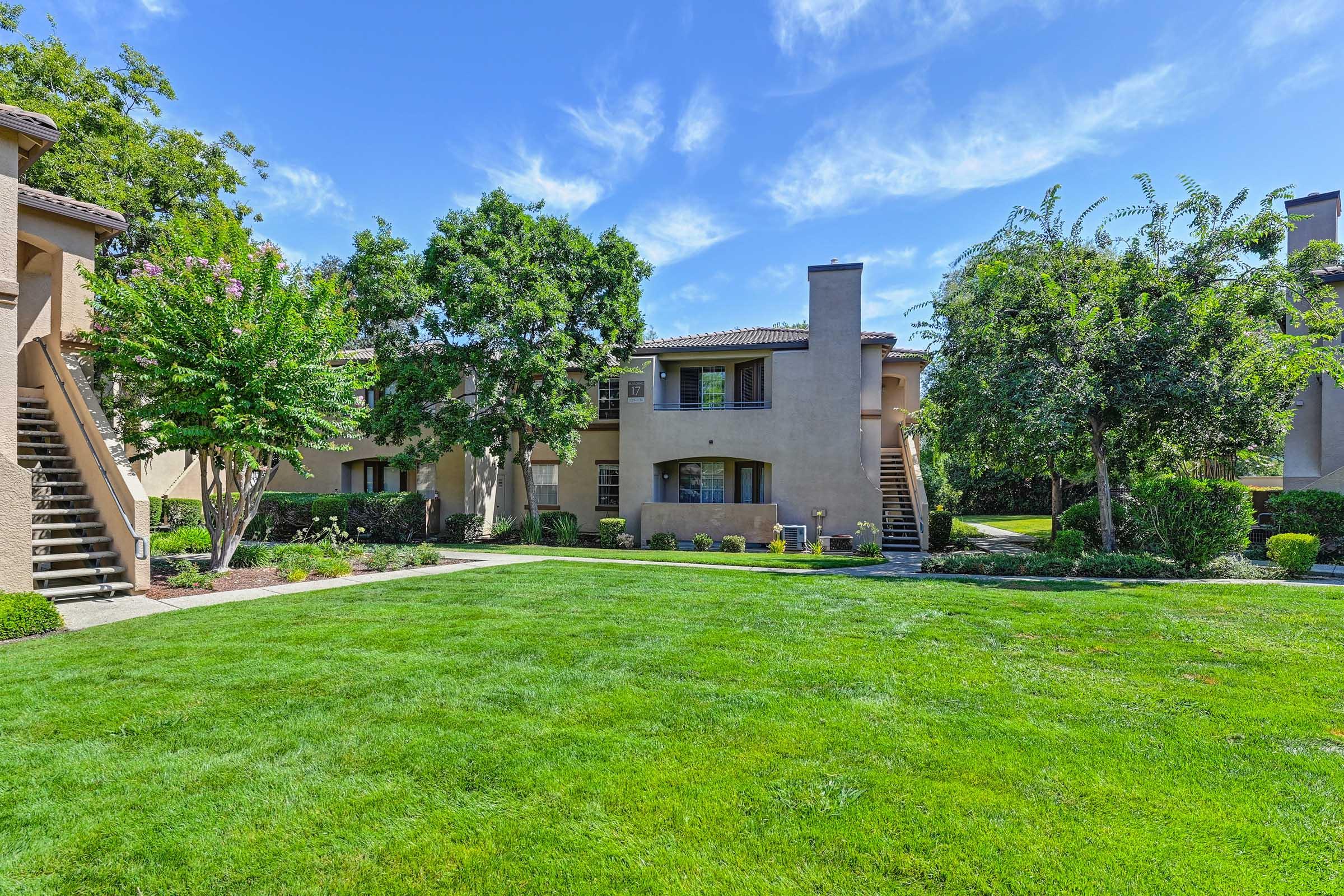 A well-maintained residential complex featuring two-story buildings surrounded by lush green grass and landscaped gardens. There are trees and shrubs adding to the aesthetic, and a clear blue sky with fluffy clouds above, creating a serene living environment.