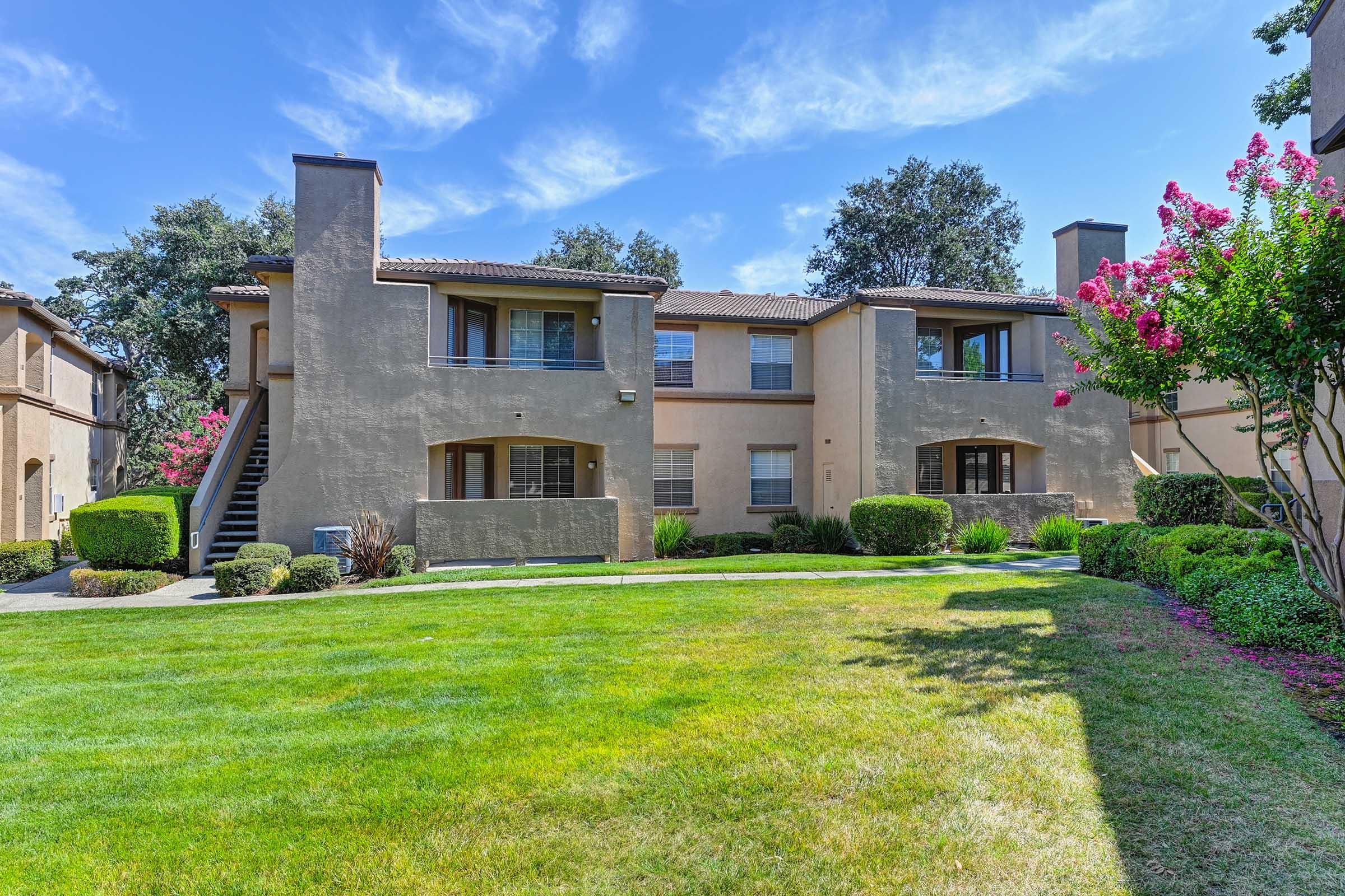 A well-maintained apartment complex with two-story buildings surrounded by lush green grass and landscaping. Flowering bushes add color to the scene under a clear blue sky, creating a pleasant and inviting atmosphere.