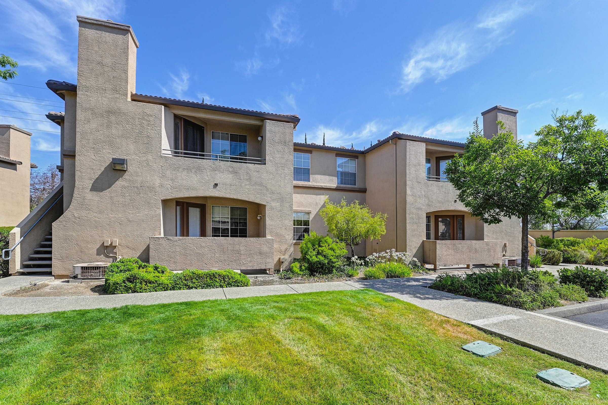 A two-story apartment building with a beige exterior, featuring balconies and landscaped grassy areas. The scene includes trees and shrubs, with a clear blue sky in the background. The walkway leads up to the entrance of the building, highlighting the well-maintained surroundings.