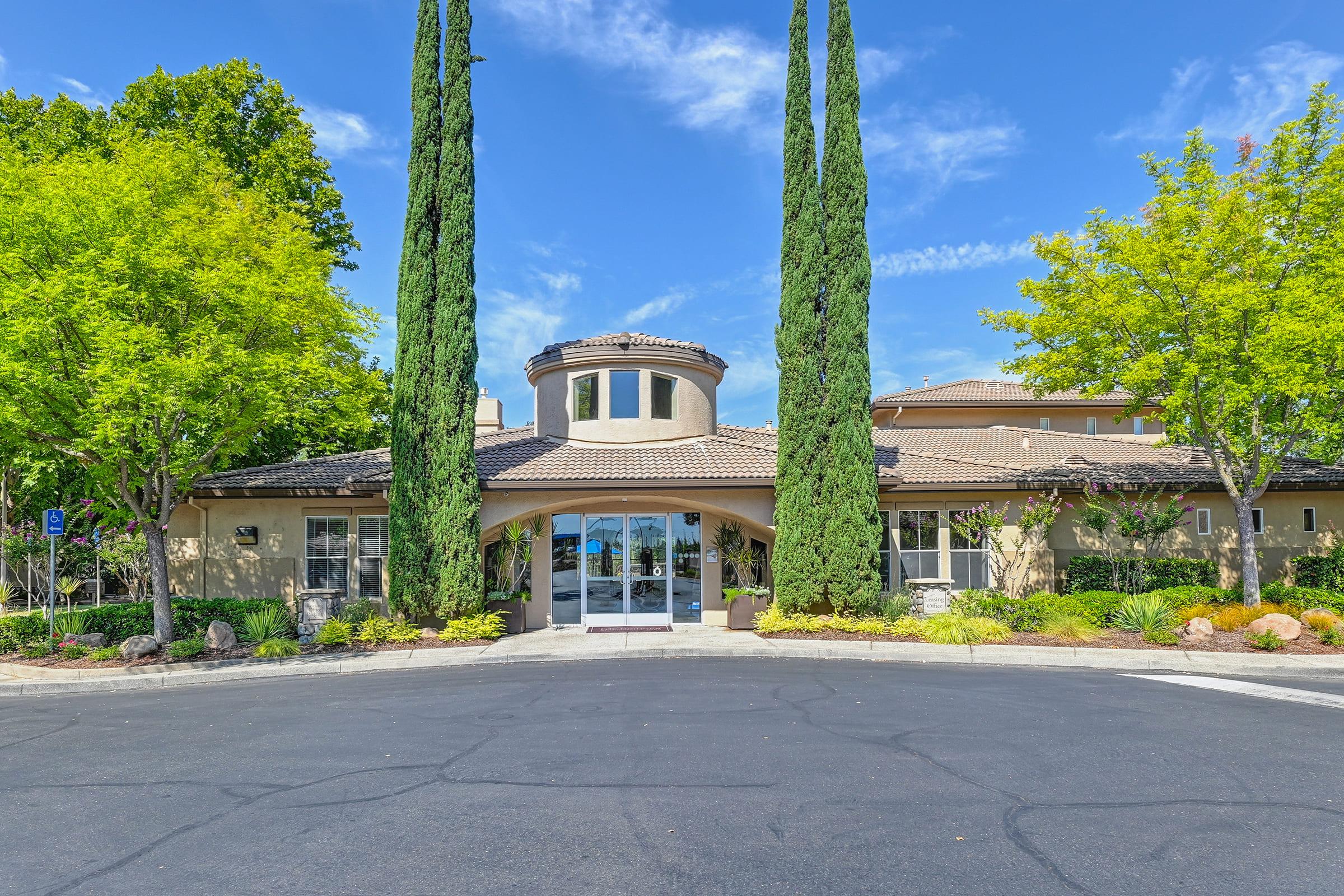 Front view of an elegant building with a rounded entrance, flanked by tall, slender cypress trees. The landscape features well-maintained greenery and colorful plants against a clear blue sky. The driveway leads up to the entrance, adding to the inviting atmosphere.