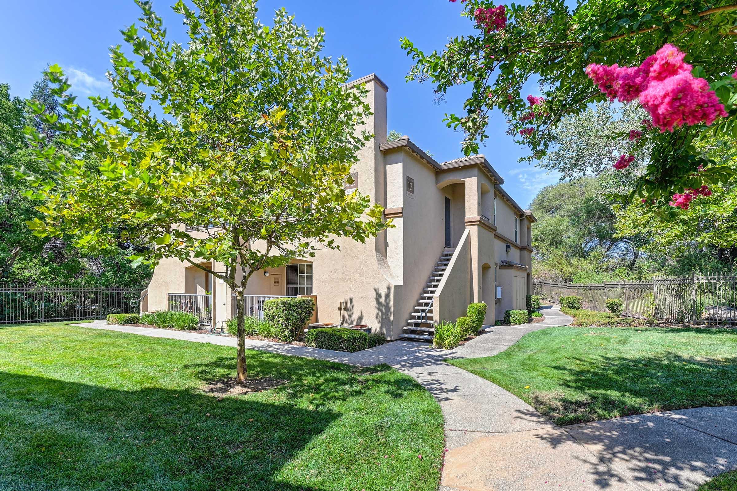 A two-story apartment building surrounded by lush greenery and colorful flowers. A pathway winds through the landscaped yard, leading to the building entrance and staircase. Sunny sky in the background enhances the inviting atmosphere of the landscape.