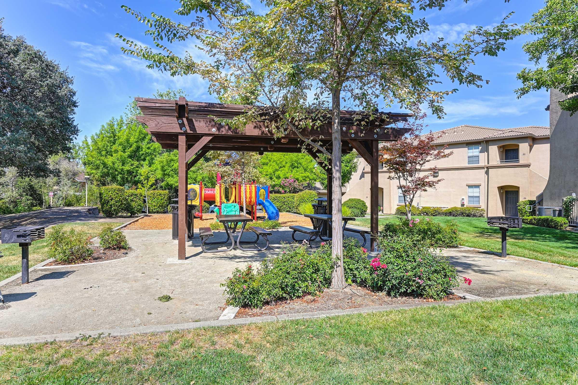 A community park with a shaded picnic area featuring a wooden pergola, tables, and barbecue grills. Nearby, there is a colorful playground with slides and climbing structures, surrounded by lush green grass and trees under a clear blue sky. Two residential buildings are visible in the background.