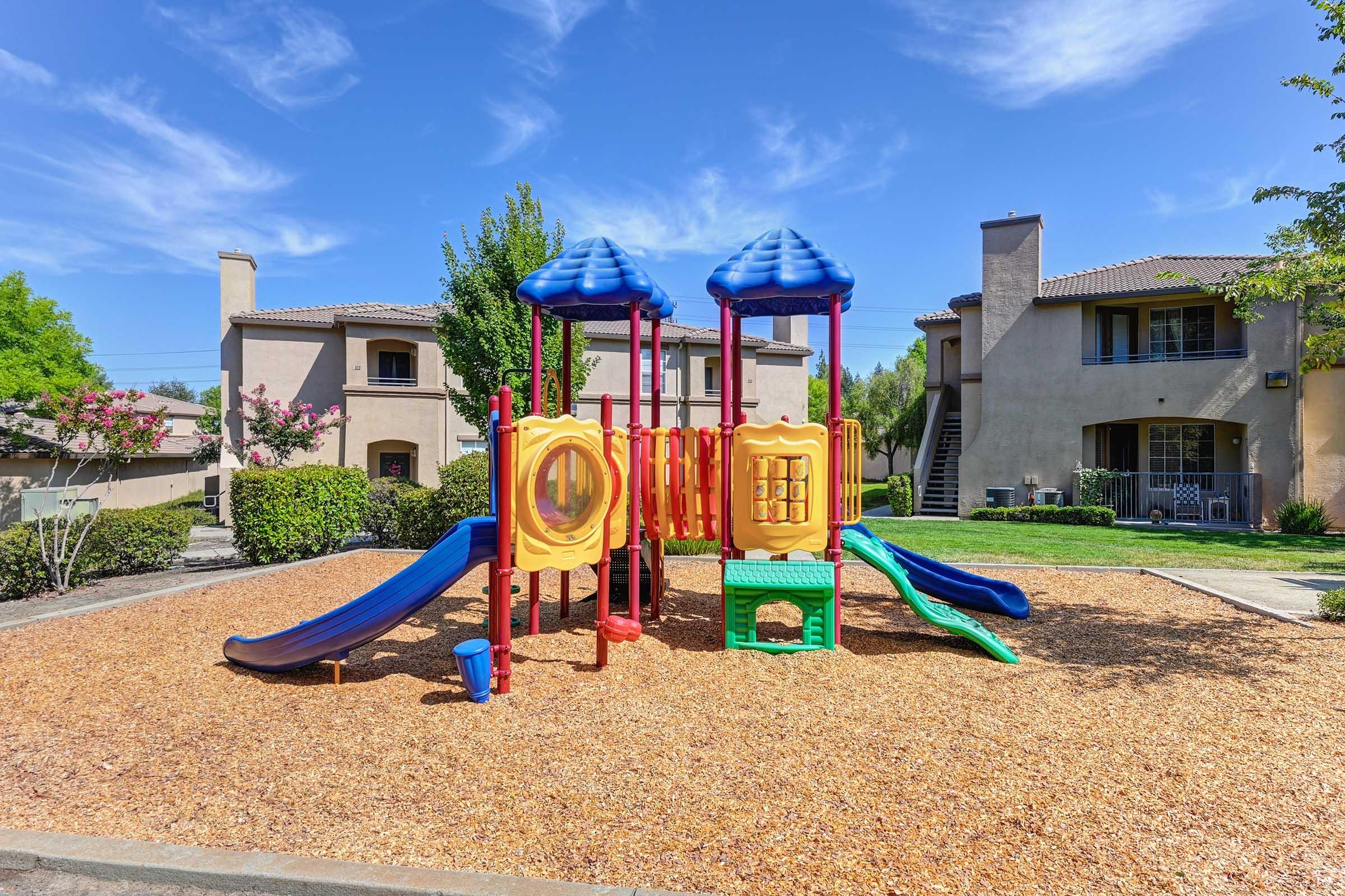 Colorful playground equipment with slides and climbing structures located on a sandy surface, surrounded by green grass and shrubs. In the background, there are multi-story residential buildings under a clear blue sky.