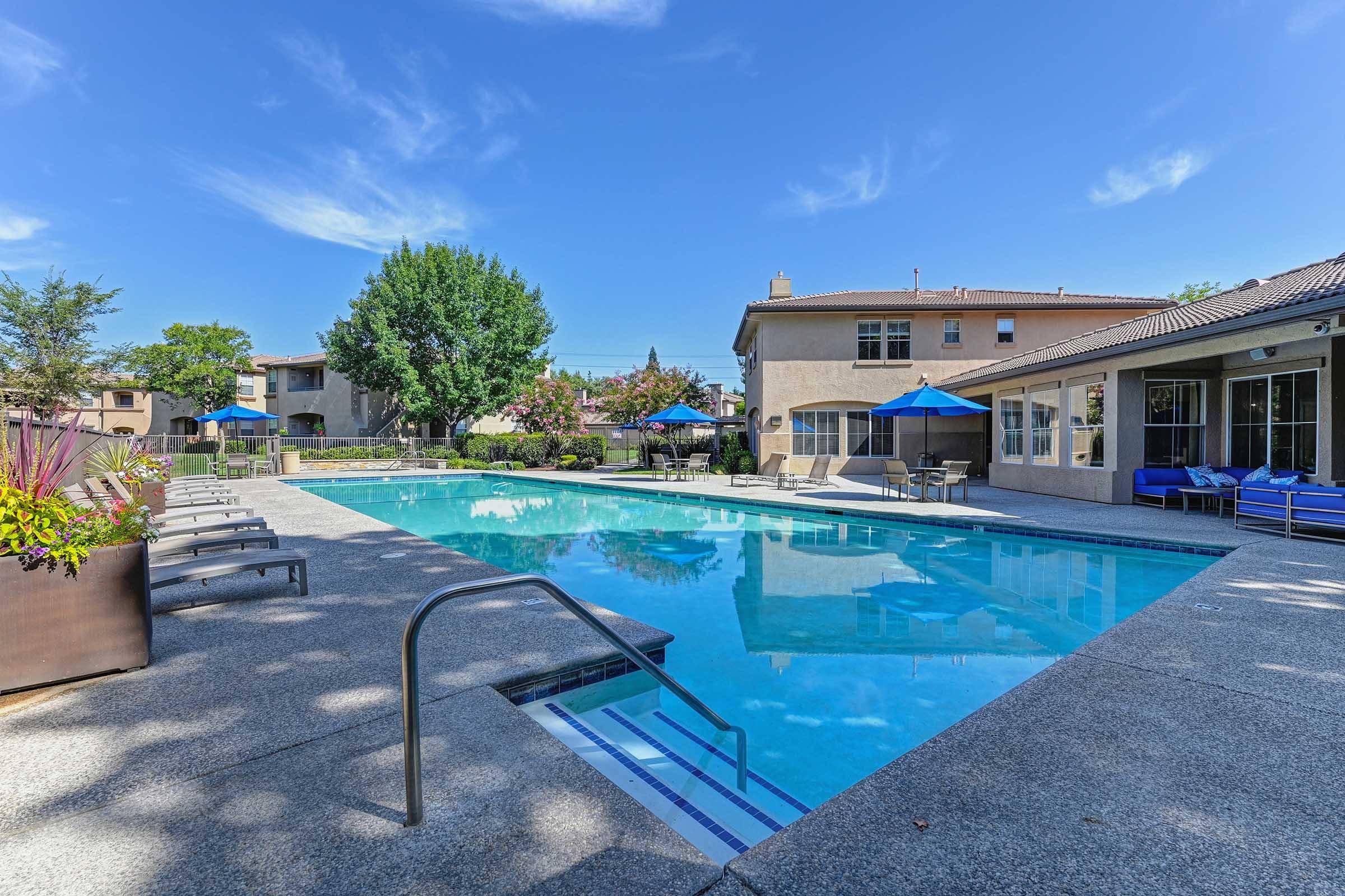 A clear blue swimming pool surrounded by lounge chairs and umbrellas, with a backdrop of residential buildings and trees under a bright blue sky. The pool area is well-maintained, inviting relaxation and leisure.