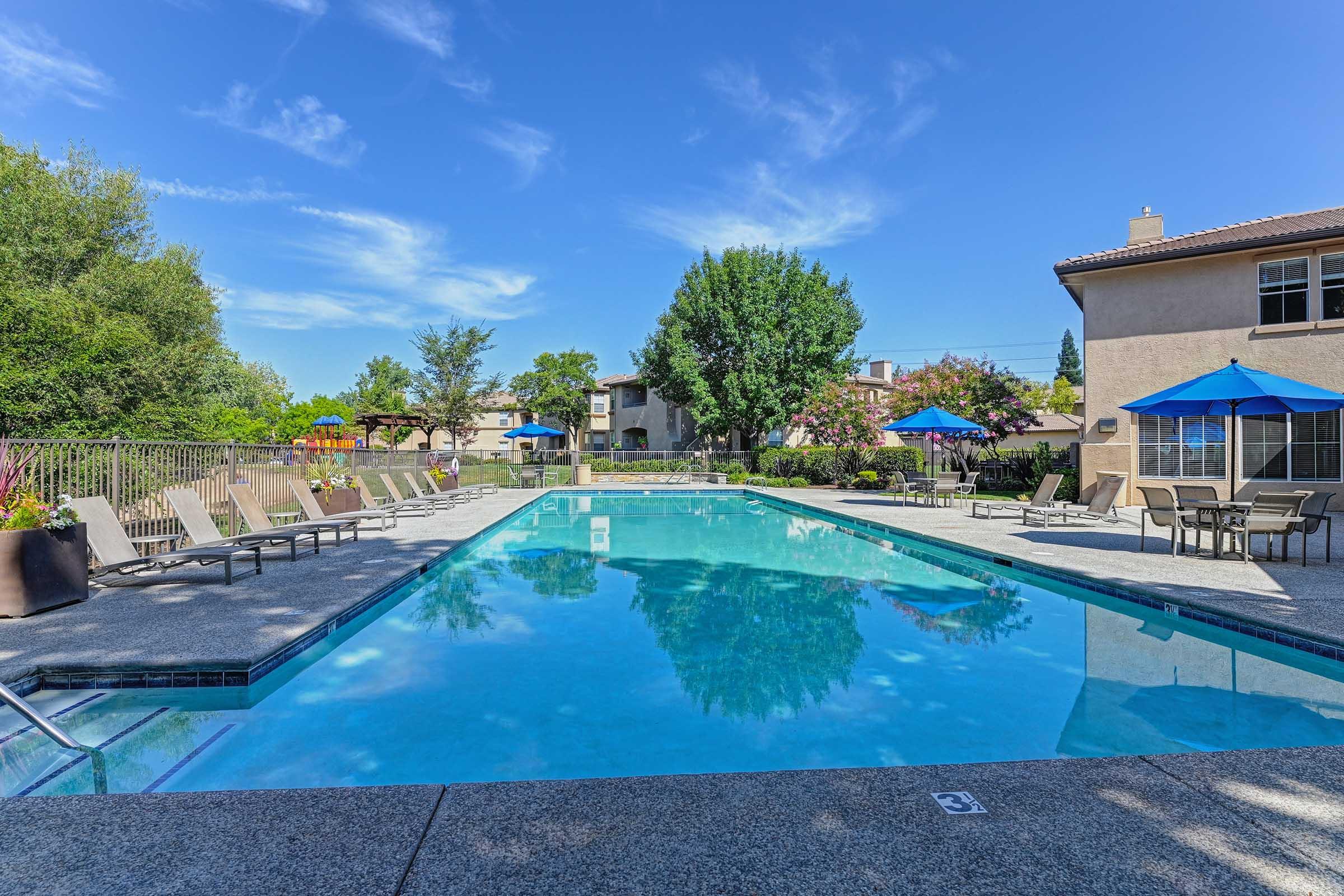 A clear blue swimming pool surrounded by lounge chairs and tables, with vibrant umbrellas providing shade. Lush green trees and colorful flowering plants frame the pool area, under a bright blue sky with a few wispy clouds. Nearby buildings can be seen in the background, adding to the serene atmosphere.