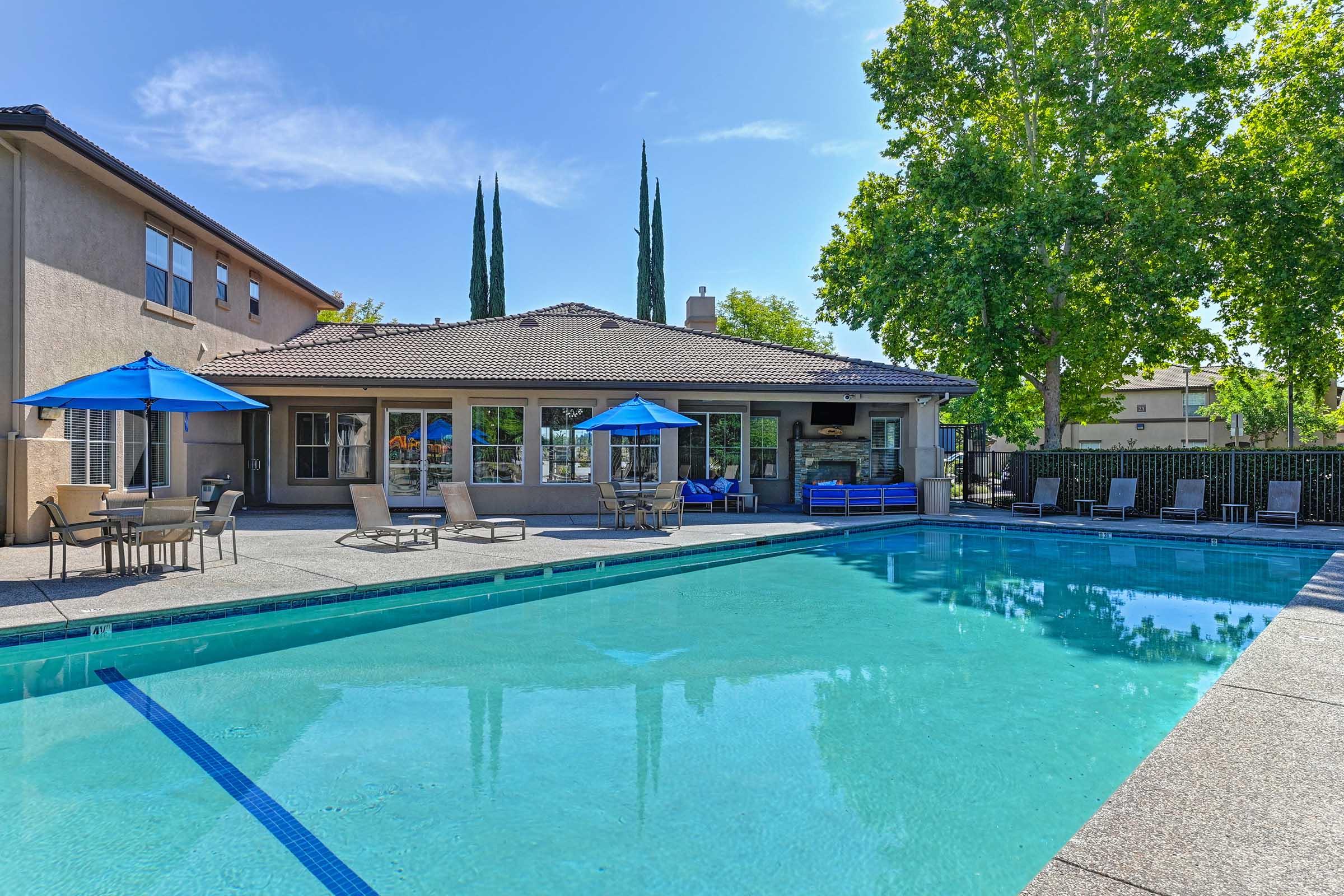 A clear blue swimming pool surrounded by lounge chairs and umbrellas. In the background, a building with large windows and a patio area is visible, along with tall trees. The sky is bright and sunny, creating a relaxing outdoor atmosphere.