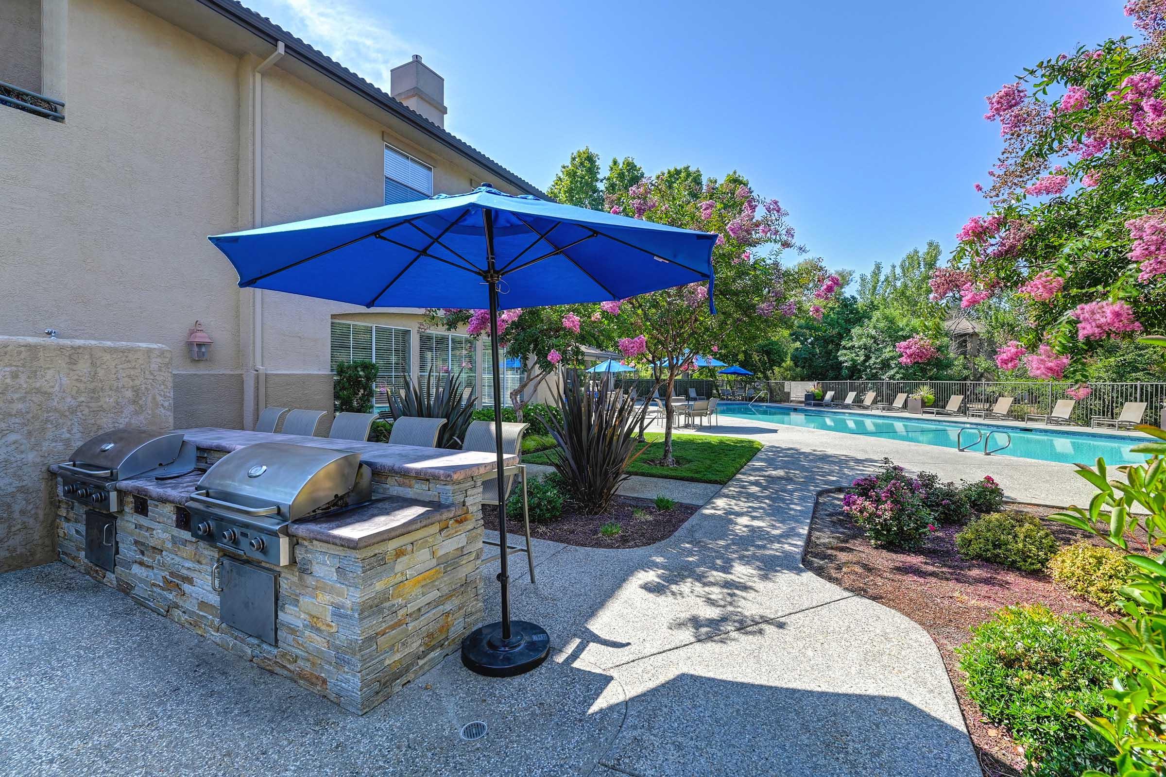 Outdoor patio area featuring a barbecue grill under a blue umbrella, surrounded by a landscaped garden with flowering trees. In the background, there is a swimming pool with lounge chairs and umbrellas, set against a clear blue sky.