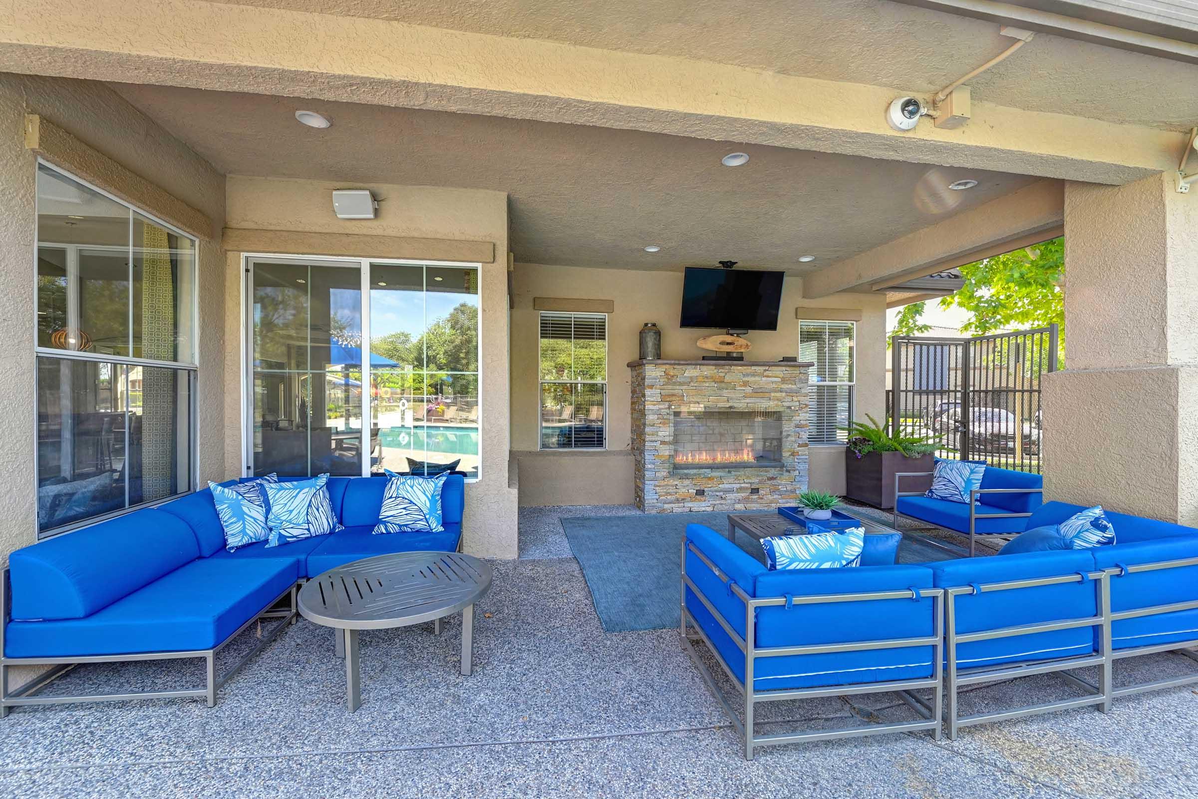 A cozy outdoor lounge area featuring blue couches with decorative pillows surrounding a circular coffee table. A stone fireplace is visible nearby, alongside large windows that overlook a swimming pool area. The space is well-lit and surrounded by greenery, creating a relaxed atmosphere.