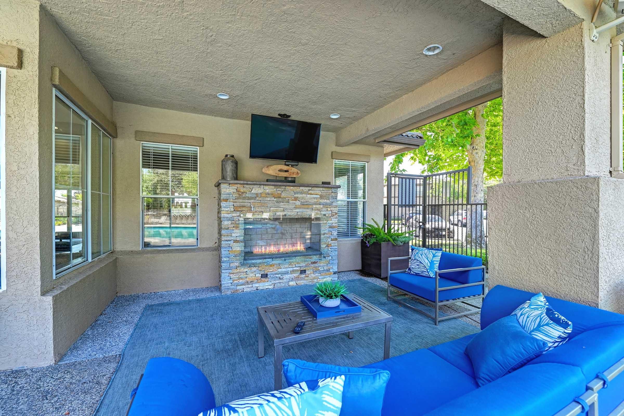 Cozy outdoor lounge with a stone fireplace, blue seating, and a TV mounted on the wall. Large windows provide natural light, and leafy trees are visible outside the enclosure. The space is designed for relaxation and socializing, featuring decorative pillows and a small table.