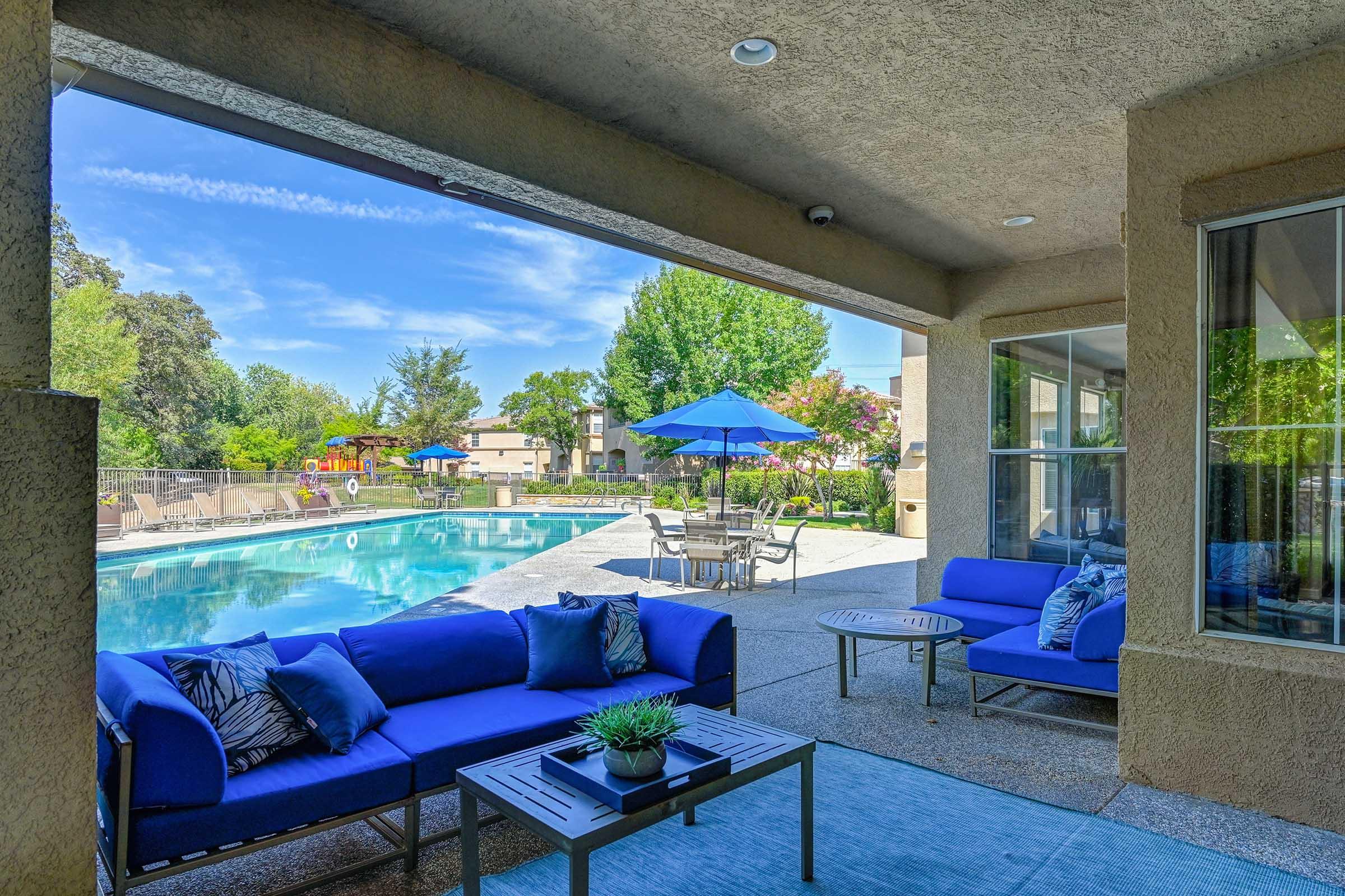 A serene outdoor lounge area featuring blue seating and a small table overlooking a clear swimming pool. The scene is framed by lush greenery and vibrant umbrellas, with sunlight casting a warm glow over the setting, inviting relaxation and enjoyment of the outdoors.