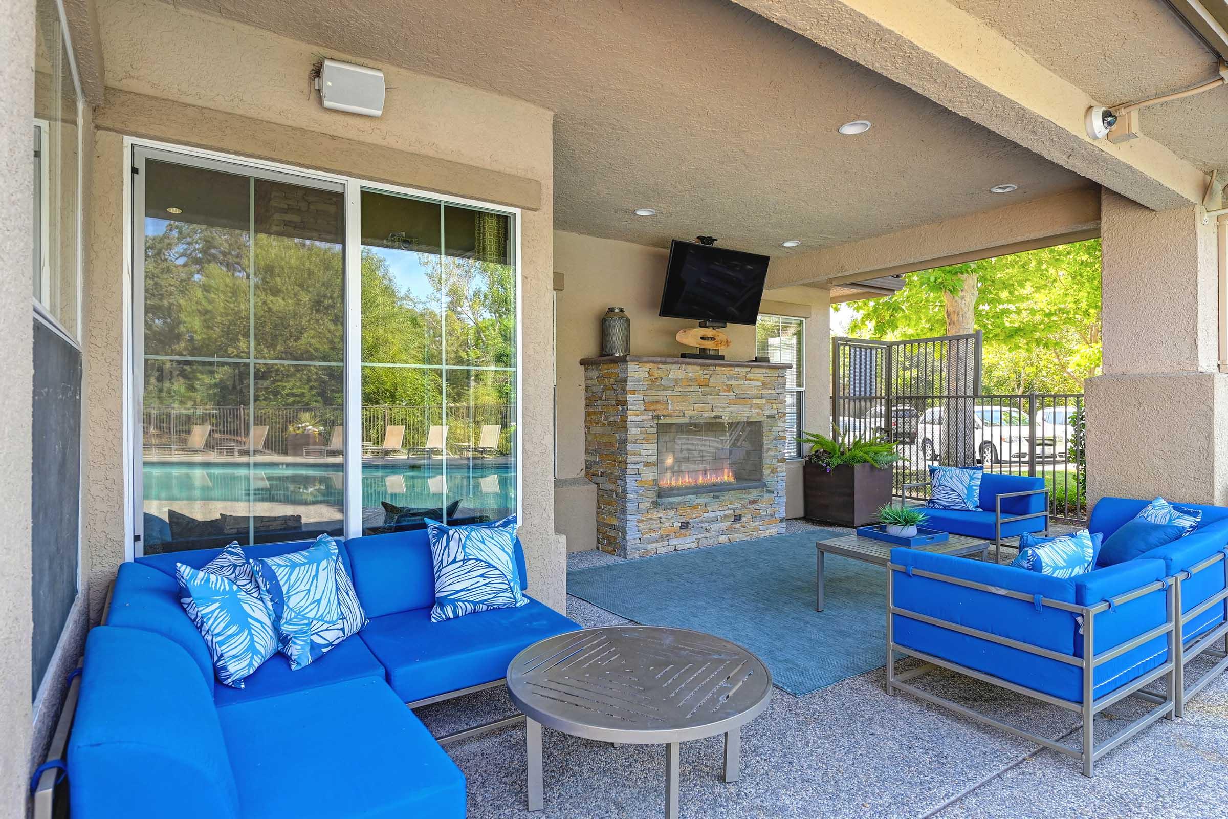 A cozy outdoor lounge area featuring blue sofas with decorative pillows, a round coffee table, and a stone fireplace. A television is mounted above the fireplace, and large windows offer a view of a pool area surrounded by greenery. The space is designed for relaxation and socializing.
