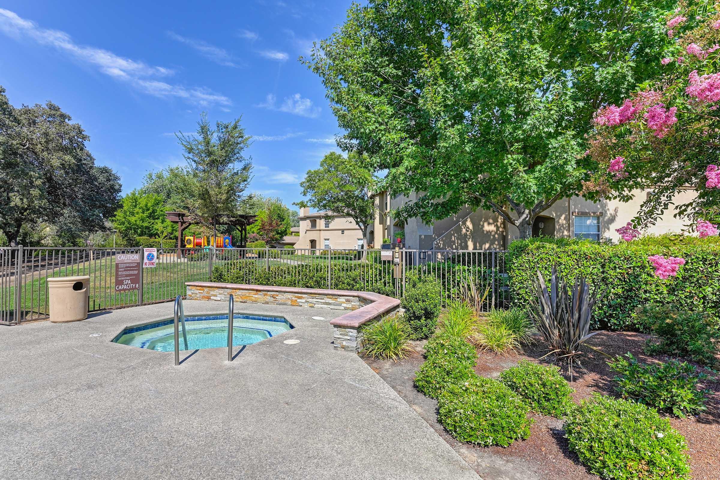 A clear blue hot tub surrounded by a landscaped area with green grass and flowering plants. In the background, there are residential buildings, trees, and a children's playground visible. The sky is bright blue with a few clouds, creating a pleasant outdoor atmosphere.