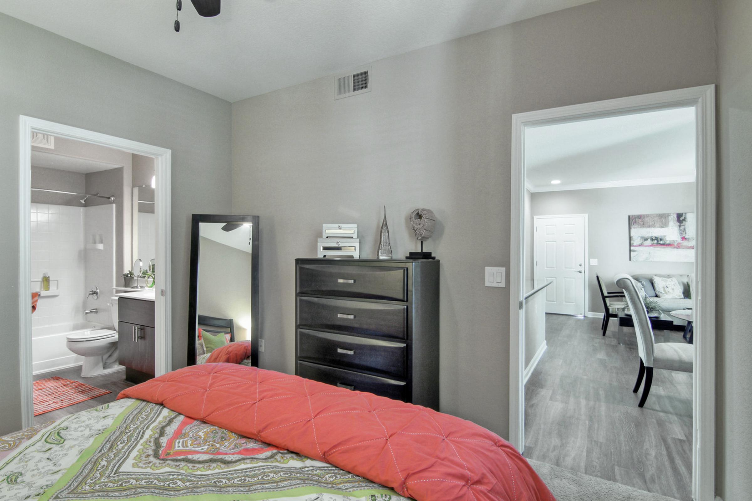 A cozy bedroom with a red and green patterned comforter on the bed. A mirror and a dark dresser are visible, along with an open door leading to a bathroom. Another doorway opens to a hallway with a dining area in view, featuring a neutral color palette and modern furnishings.