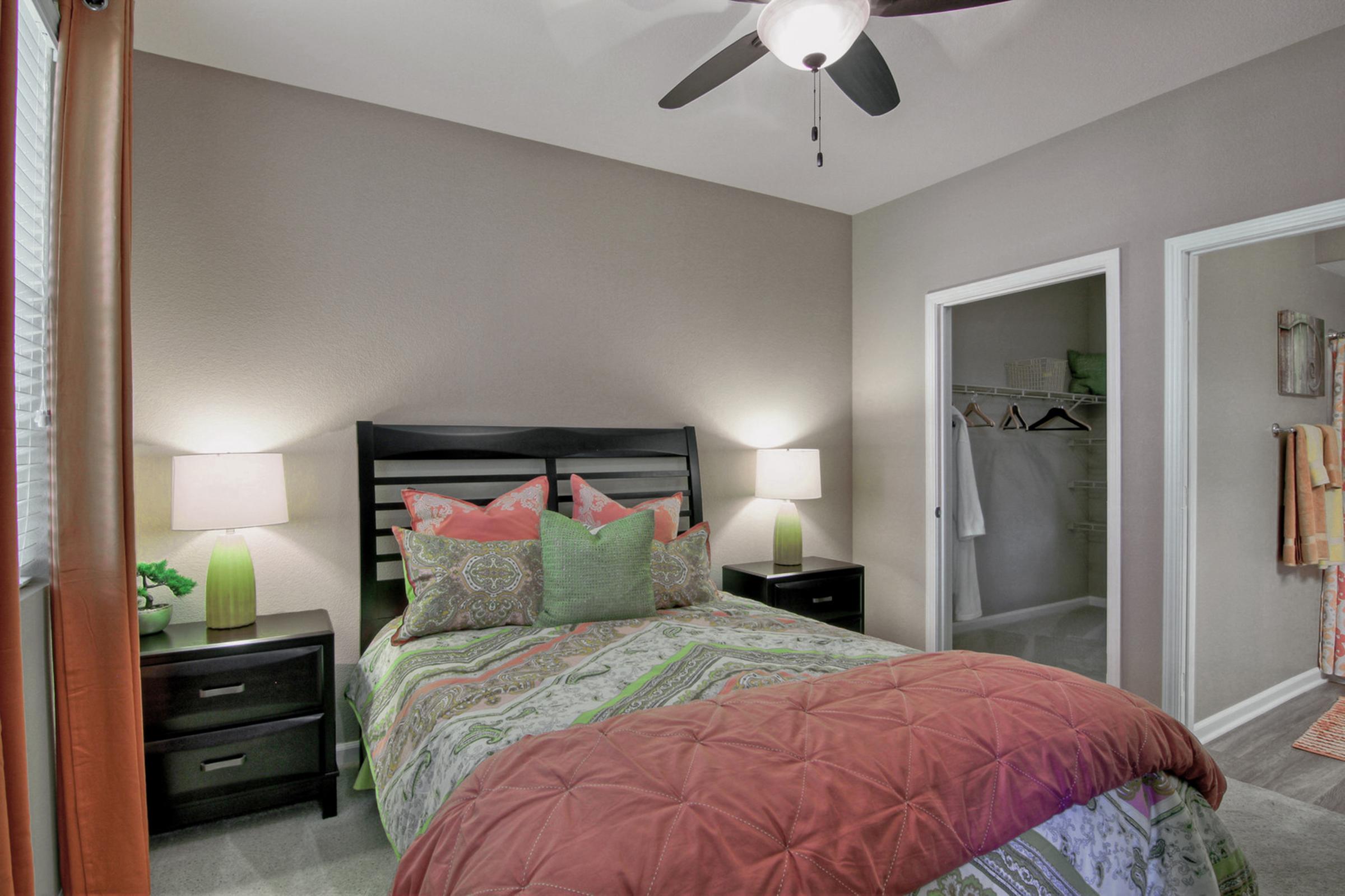 A stylish bedroom featuring a black wooden bed with colorful patterned bedding and several decorative pillows. Two bedside lamps illuminate the space, and there is a green plant on a nightstand. A closet is visible in the background, along with an adjoining bathroom area, creating a cozy and inviting atmosphere