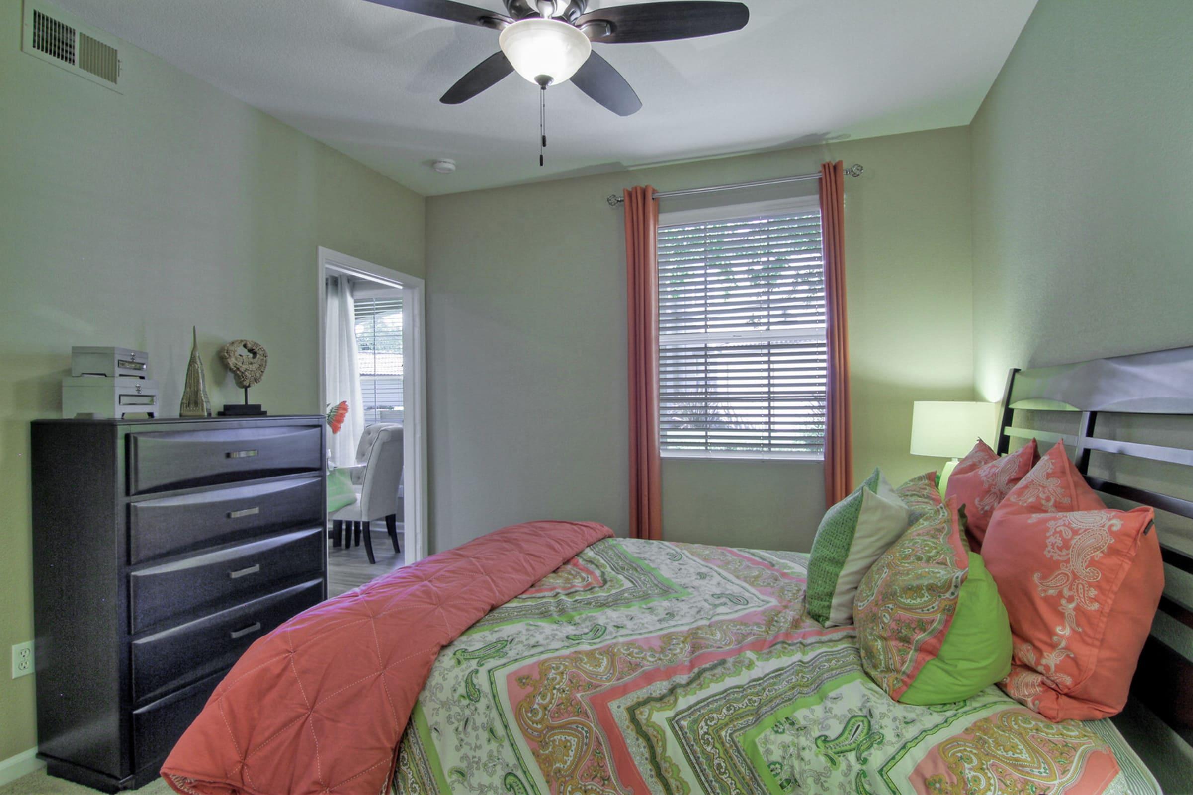 A cozy bedroom featuring a large bed with colorful bedding in shades of green and coral. There is a ceiling fan, a bedside lamp, and a black dresser. Natural light filters through a window with blinds, and an adjoining room is visible in the background with a dining area.