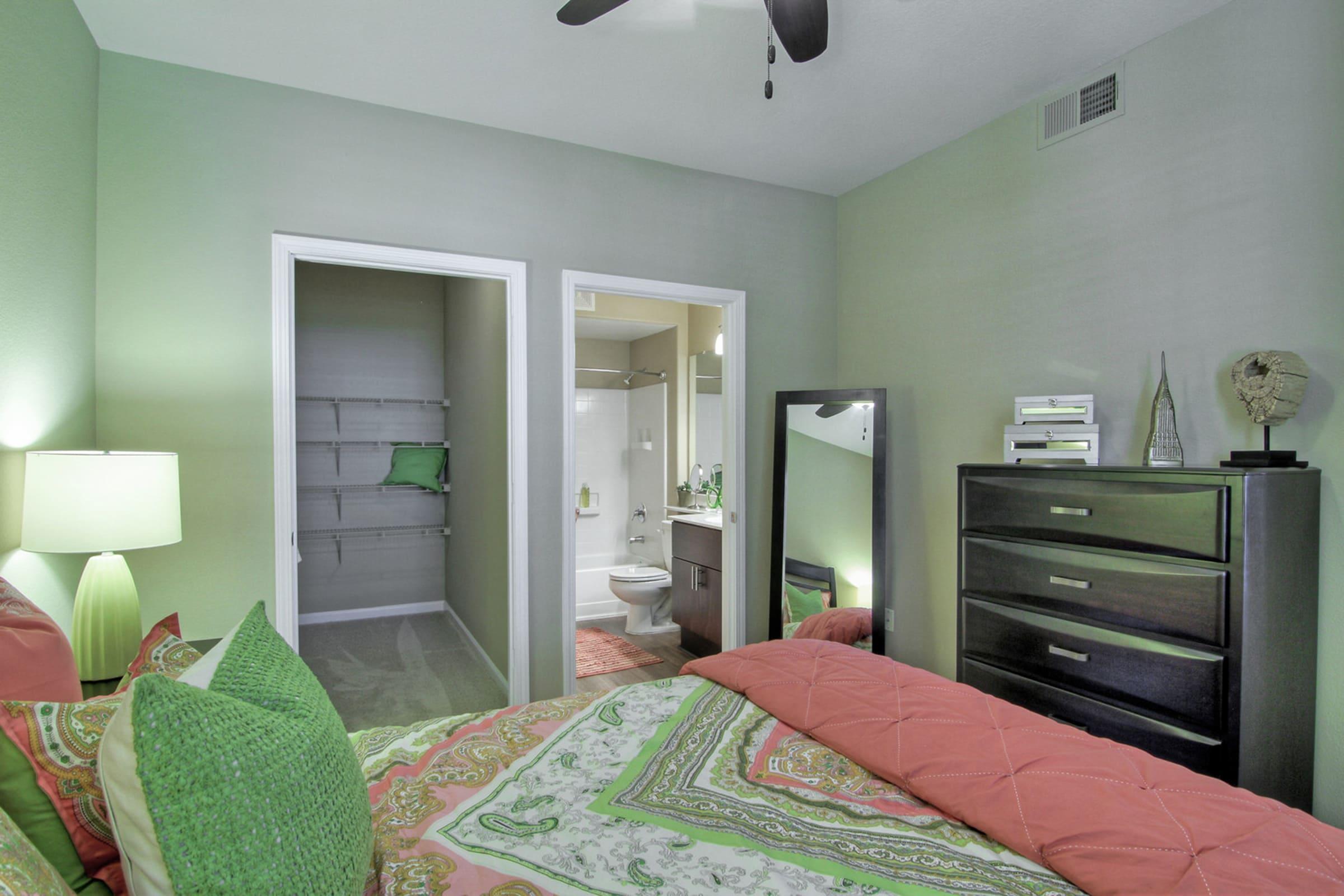 A cozy bedroom featuring green walls, a bed with colorful bedding, and a bedside lamp. There is a large mirror and a dark wood dresser. A door leads to a small bathroom area, and another door opens to a closet with shelves. The room has a modern and inviting atmosphere.
