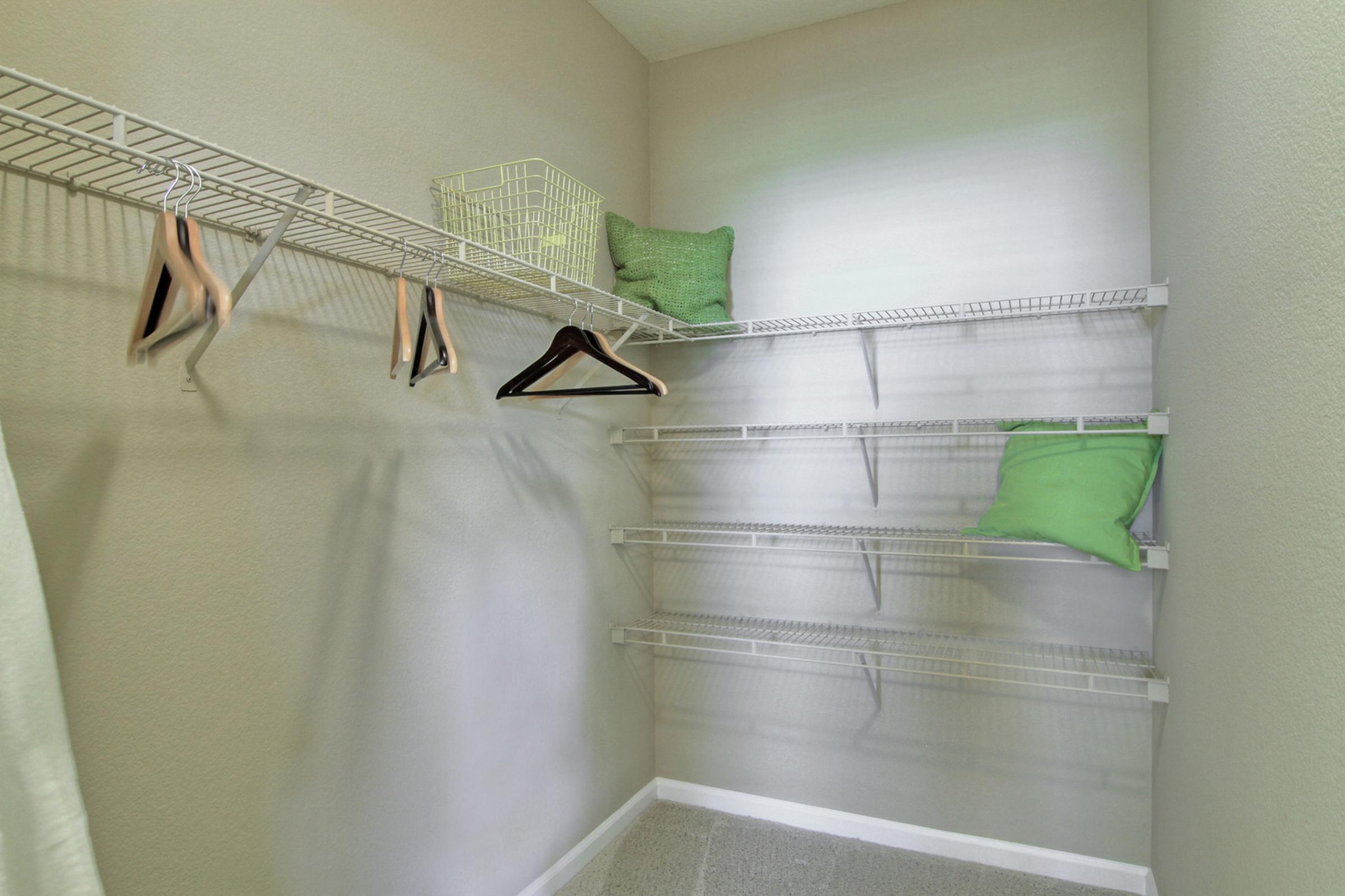 A well-organized, empty closet featuring white wire shelving, a few green decorative pillows, and a couple of wooden and plastic hangers. The walls are painted in a light tone, creating a clean and spacious appearance.