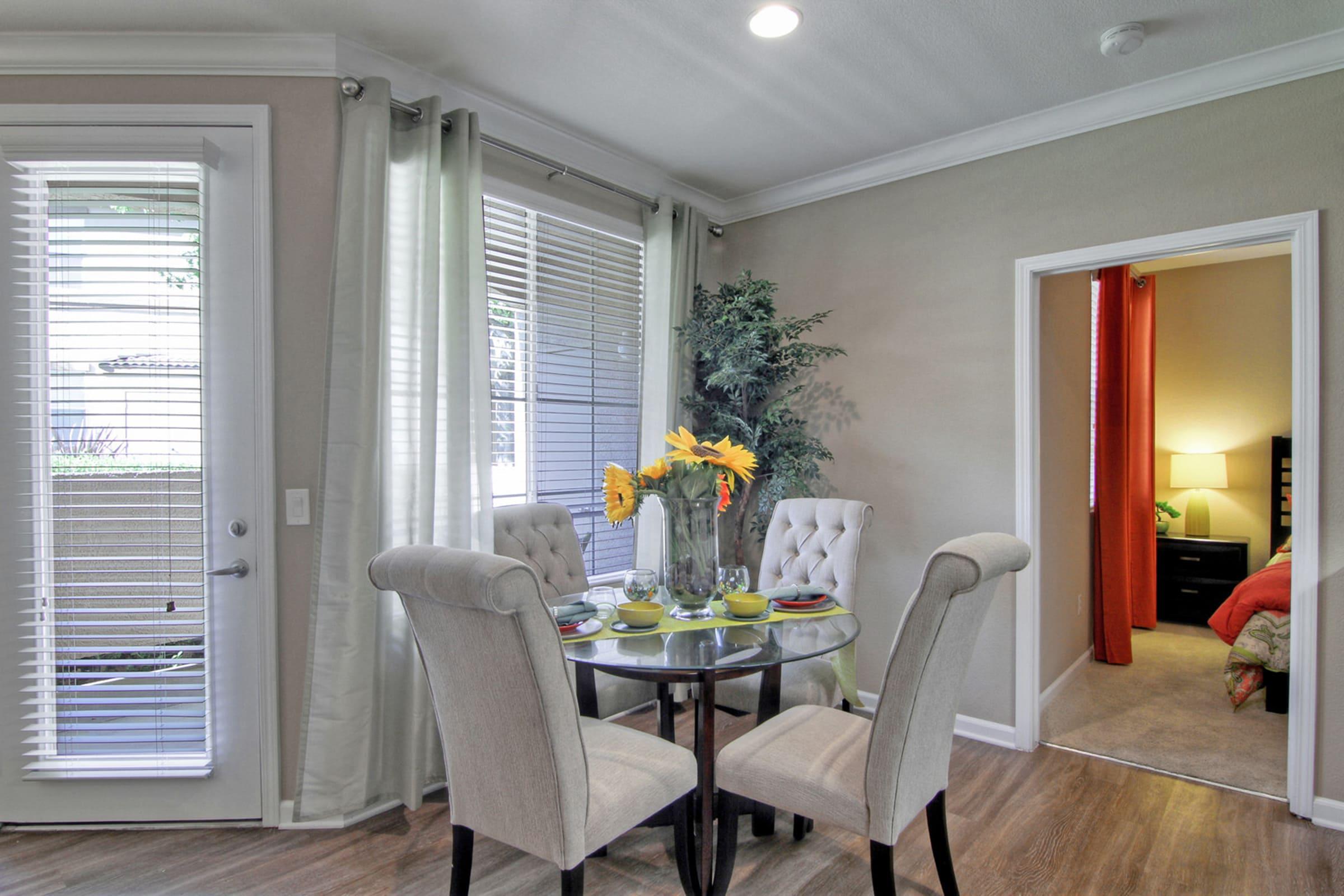 A cozy dining area featuring a round glass table set for a meal with plates and flowers. Two upholstered chairs surround the table. Bright curtains frame the window, allowing natural light to enter. In the background, a doorway leads to a bedroom with warm-toned decor.