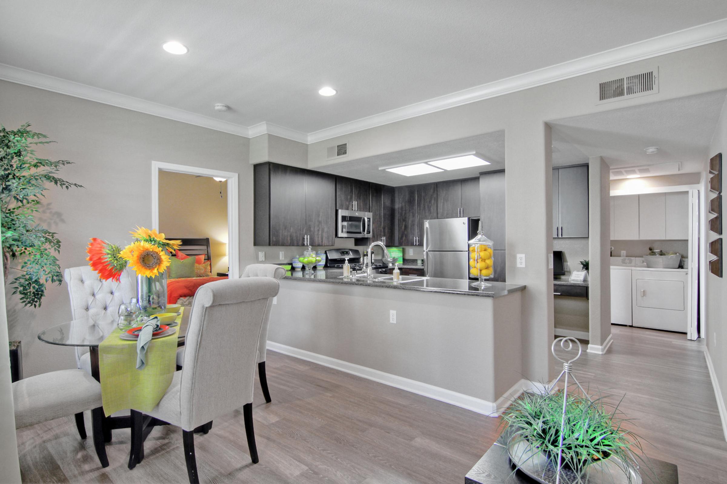 Bright and modern kitchen and dining area featuring a table set for four with flowers, sleek black cabinets, stainless steel appliances, and a bar counter. Natural light fills the space, enhancing the open layout. A cozy living area is visible in the background, adding warmth to the home.