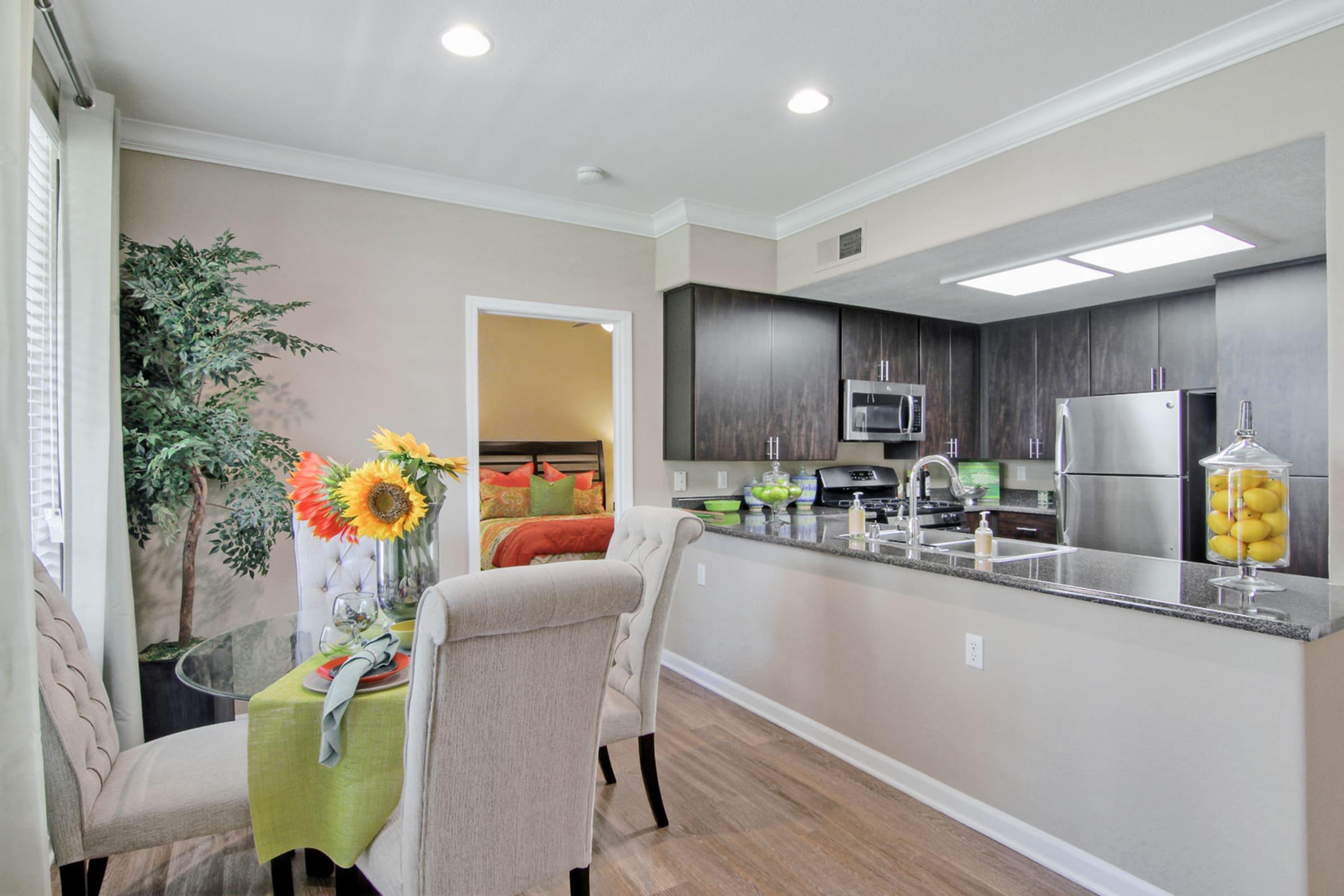Modern kitchen with dark cabinetry and stainless steel appliances. A round table with a vase of sunflowers is set for dining, accompanied by a stylish chair. A decorative plant is visible in the corner, and the space is illuminated by overhead lights, creating a warm and inviting atmosphere.