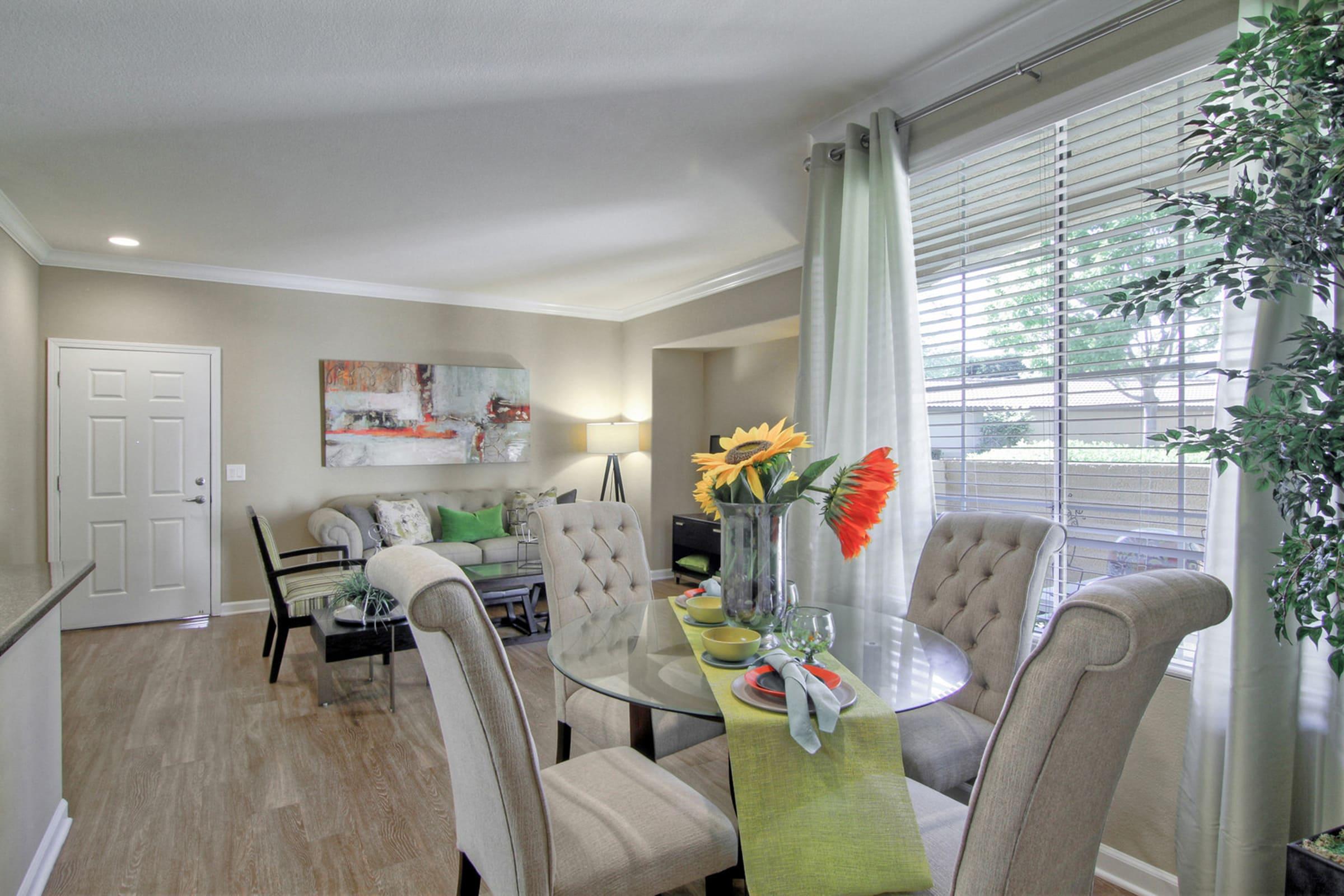 A modern dining area featuring a round glass table set with colorful dishes and a vase of flowers. Stylish chairs surround the table, and natural light streams through a window with sheer curtains. A decorative painting hangs on the wall, and indoor plants add greenery to the space.