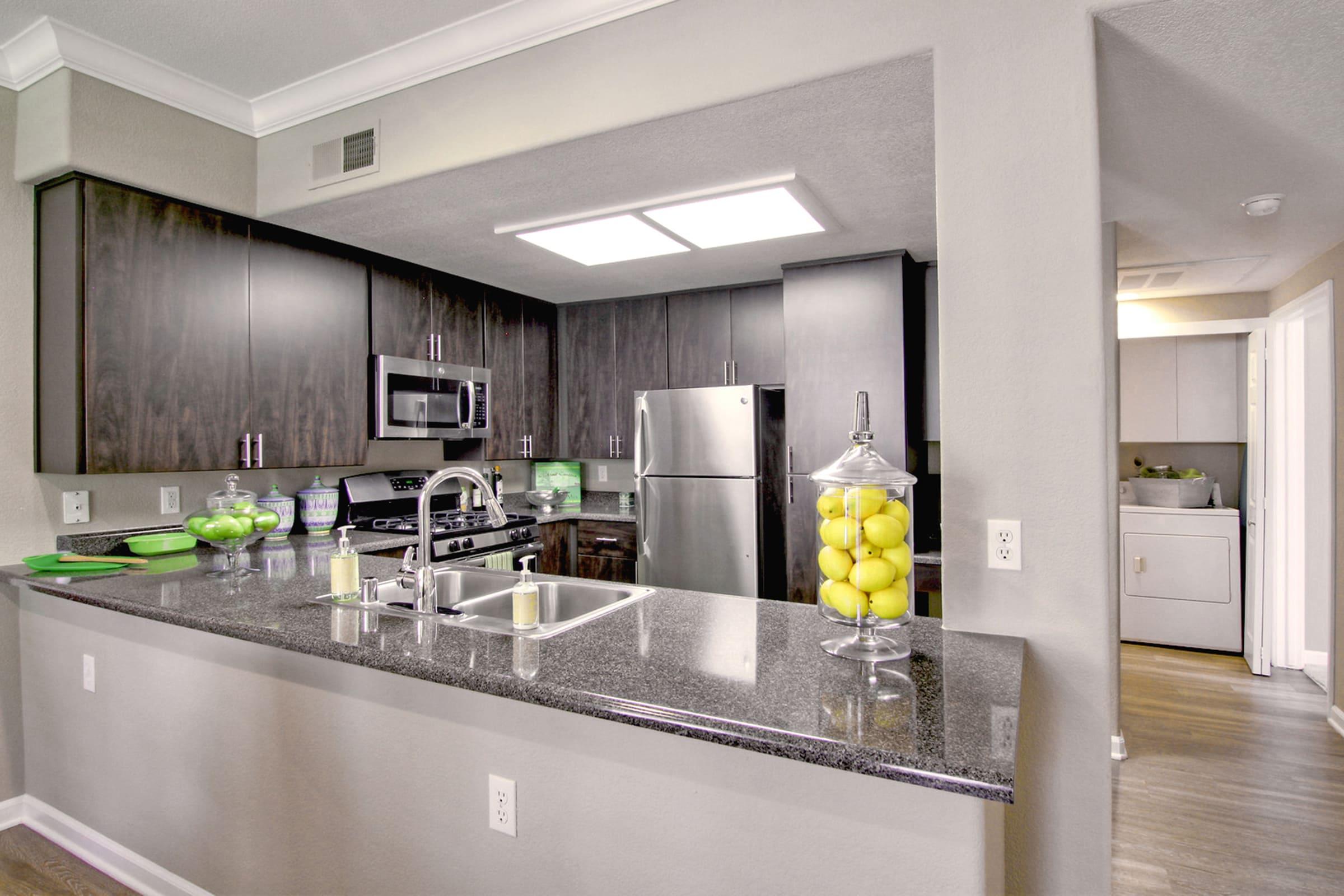 Modern kitchen featuring dark cabinetry, stainless steel appliances, and a granite countertop. A glass container filled with lemons is prominently displayed, along with green kitchen accents. The open layout leads to a visible laundry area. Bright lighting enhances the contemporary atmosphere.