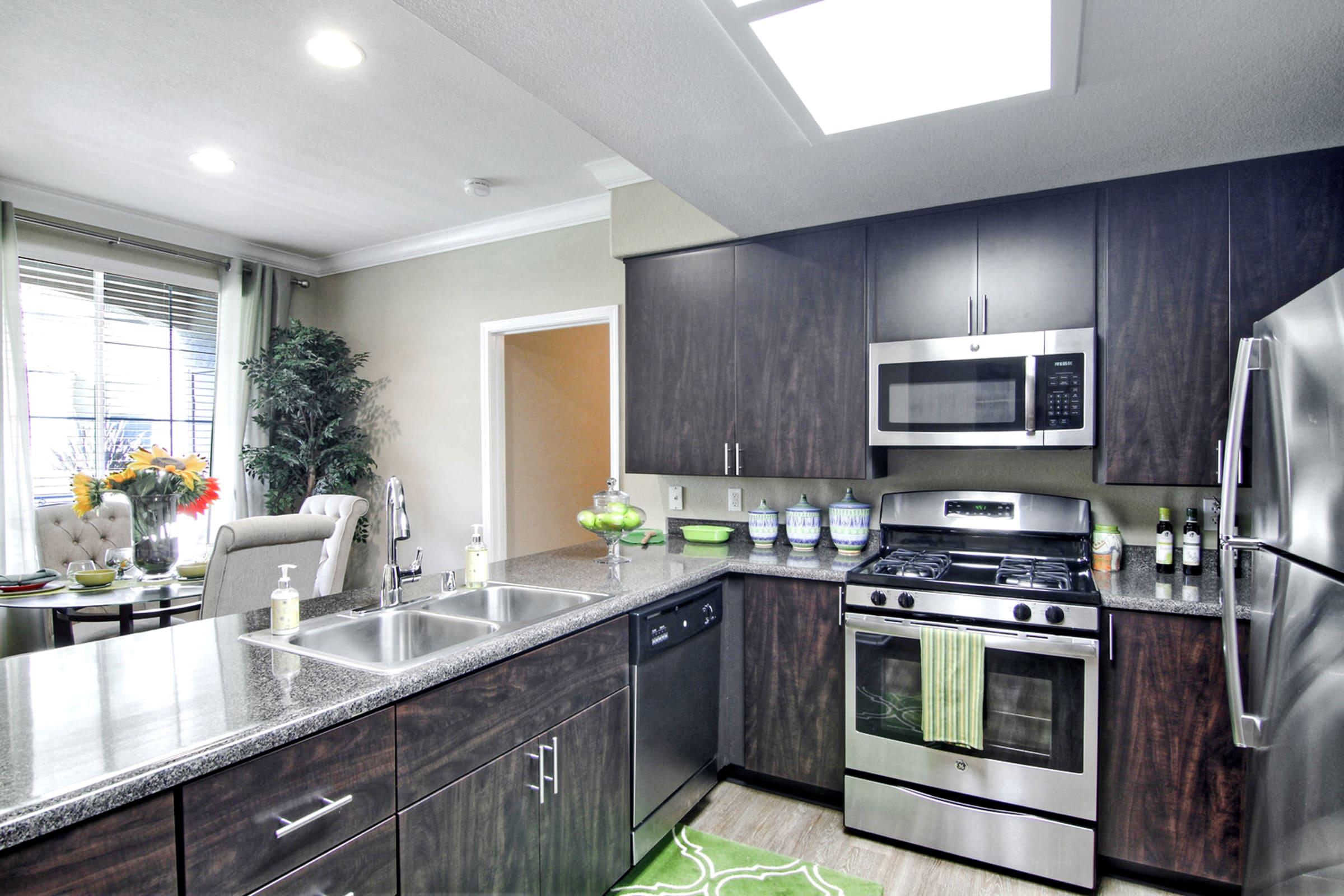 Modern kitchen featuring dark wood cabinetry, stainless steel appliances, and a gray countertop. Includes a double sink, a gas stove, and decorative items on the counters. A dining area with a table and chairs is visible in the background, along with natural light coming through the windows.