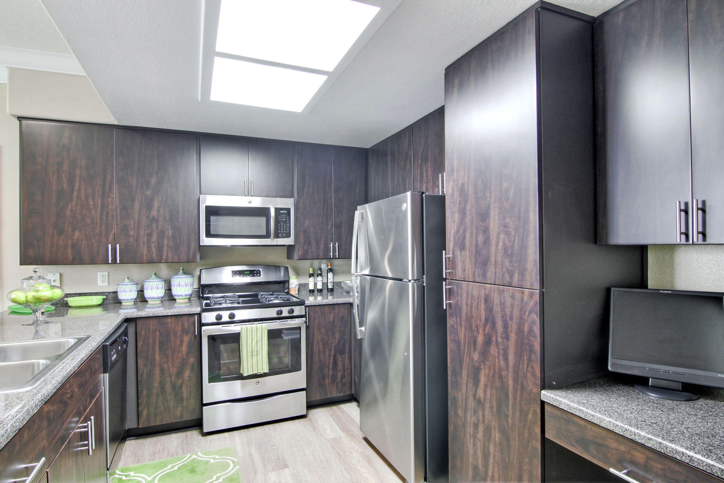 Modern kitchen featuring dark wood cabinets, stainless steel appliances including a microwave, gas stove, and refrigerator. The countertops are grey with a textured finish, and a small computer monitor sits on the counter. Bright ceiling lights illuminate the space, creating a welcoming atmosphere.
