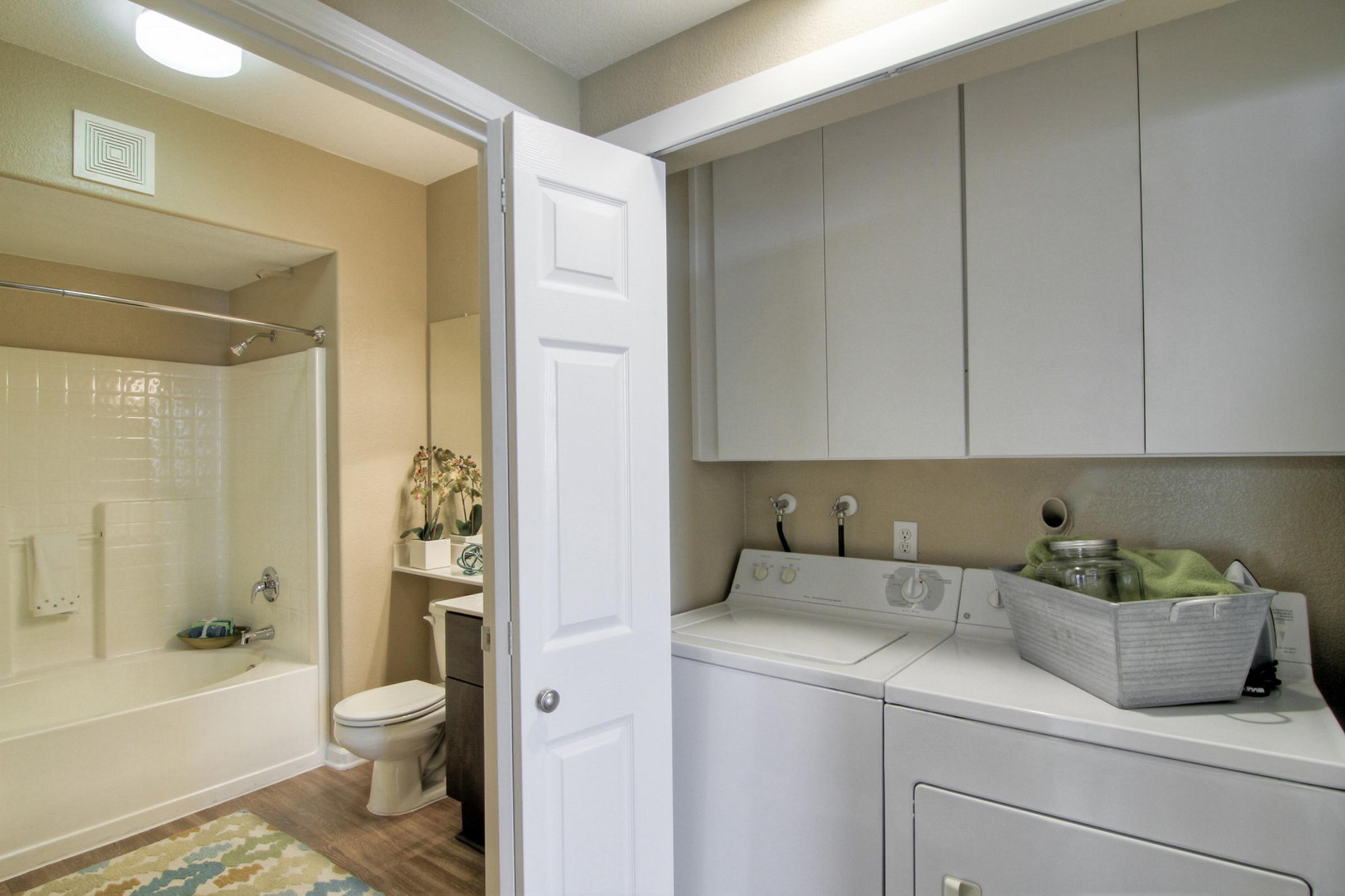 A laundry area with a washer and dryer next to a bathroom. The bathroom features a bathtub and a toilet. The space is bright with neutral-colored walls, and decorative items are placed on the countertop. A woven basket holding towels or linens sits on top of the dryer.