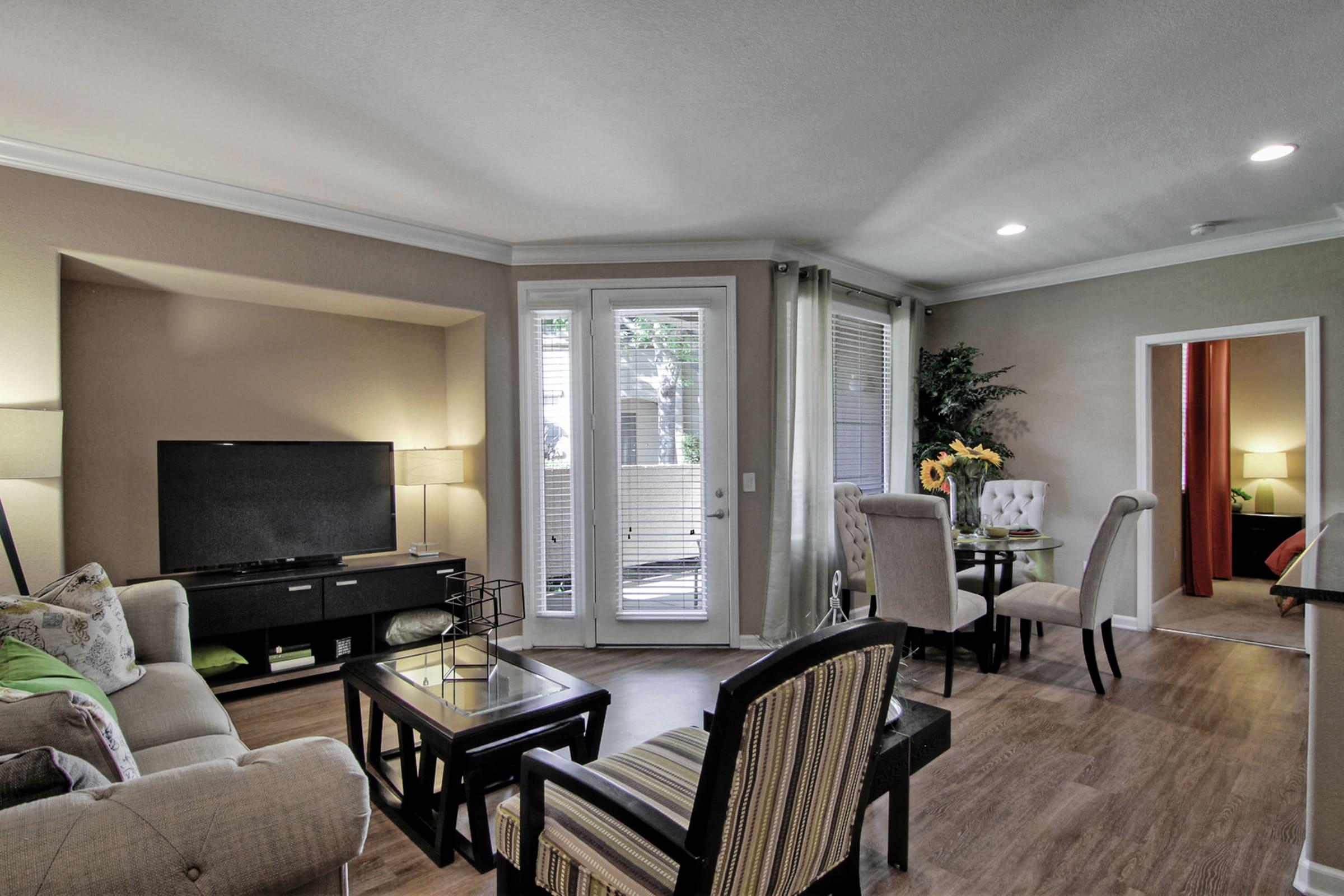 A cozy living room featuring a beige sofa, a black coffee table, and a television on a stand. Natural light enters through large windows with sheer curtains. A small dining area with a round table and chairs is visible, along with decorative elements like flowers. A doorway leads to another room.