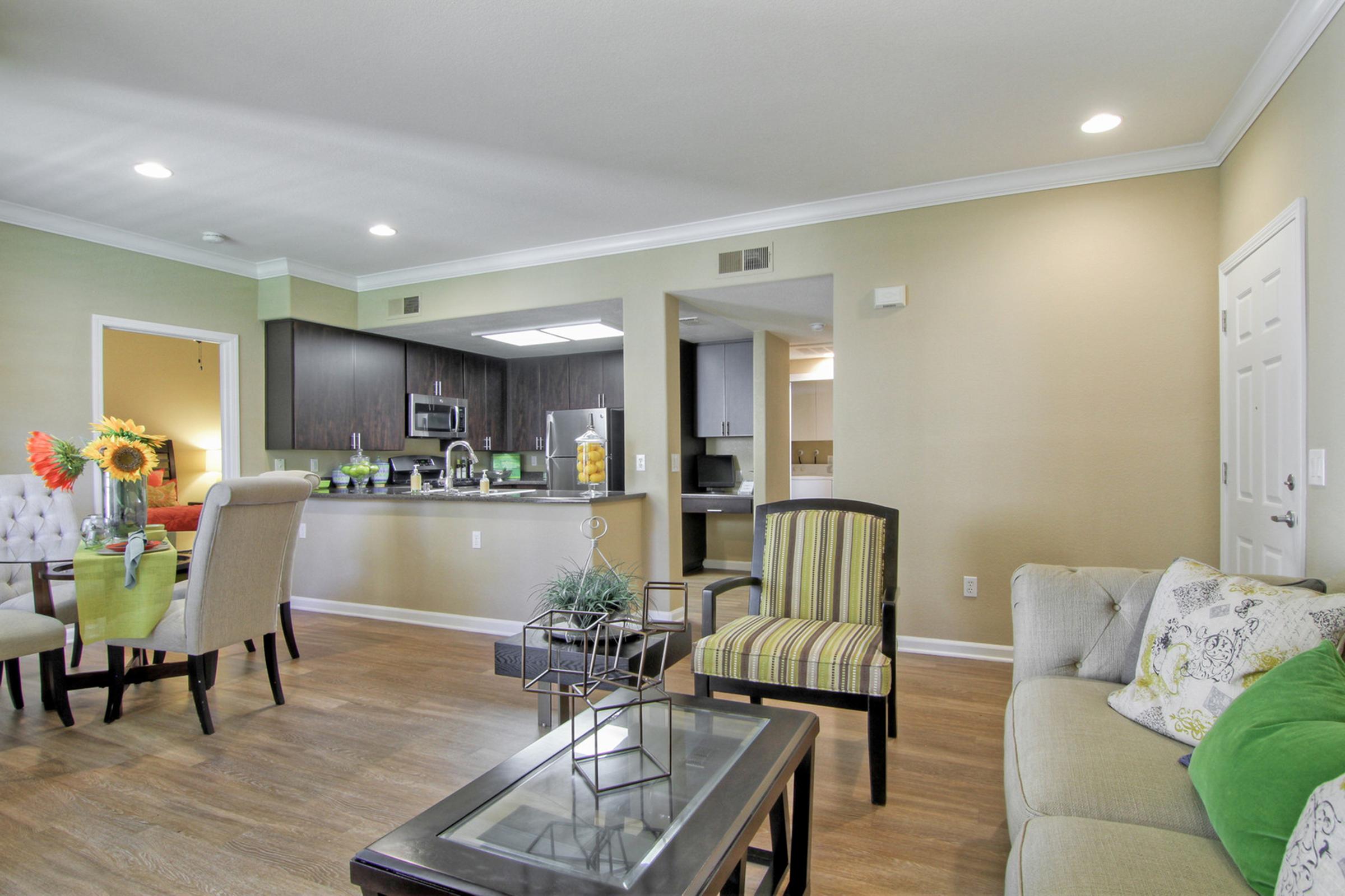 A modern living room and kitchen area featuring a cozy sofa with decorative pillows, a glass coffee table, dining chairs, and a dining table with a vibrant centerpiece of flowers. The walls are painted light green, and the kitchen is equipped with dark cabinets and stainless steel appliances