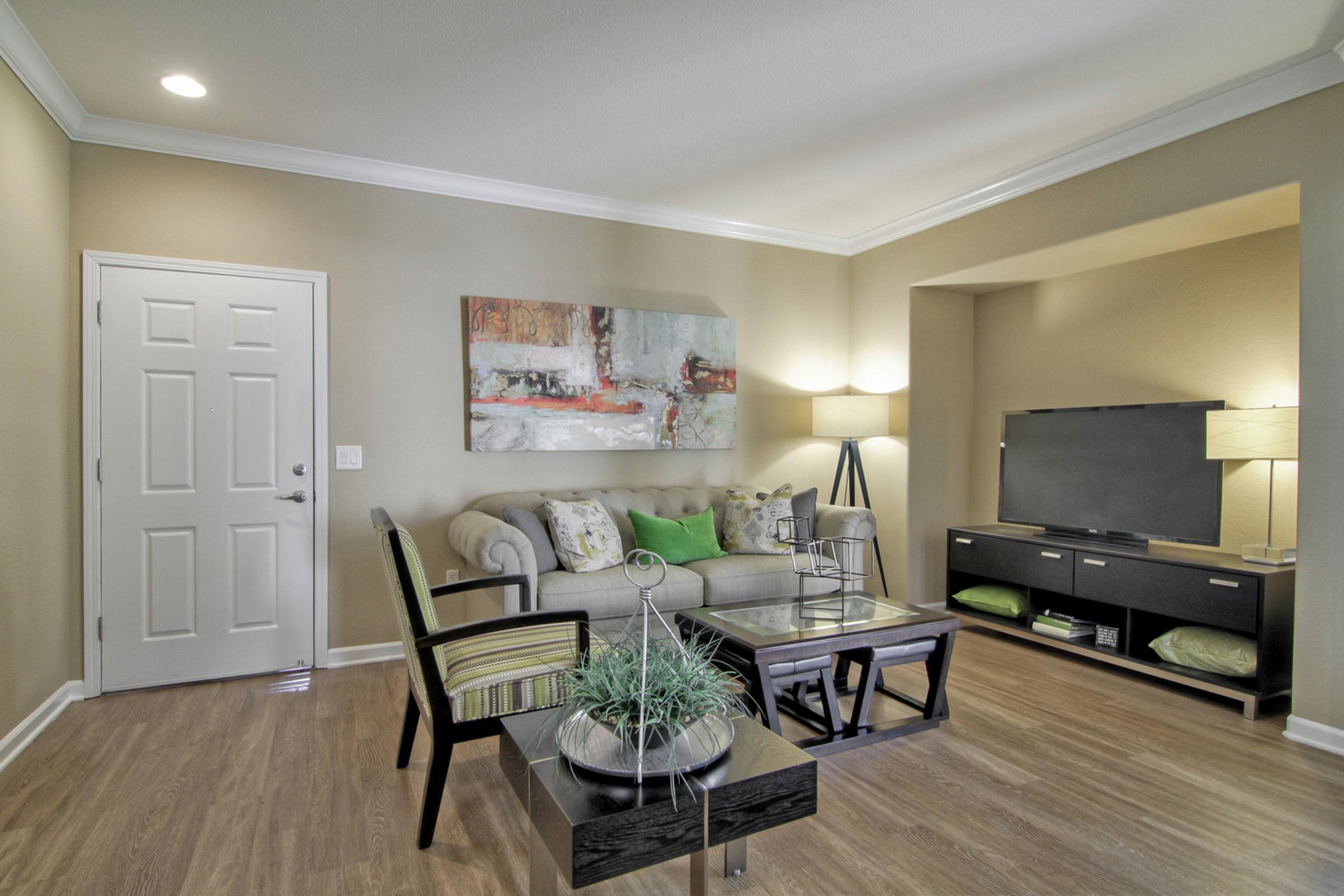 Modern living room featuring a beige couch with colorful pillows, a glass coffee table on a light hardwood floor, and a TV unit. There are two lamps providing ambient lighting, and a large piece of abstract art hangs on the wall. A decorative plant is placed on the coffee table, enhancing the decor.