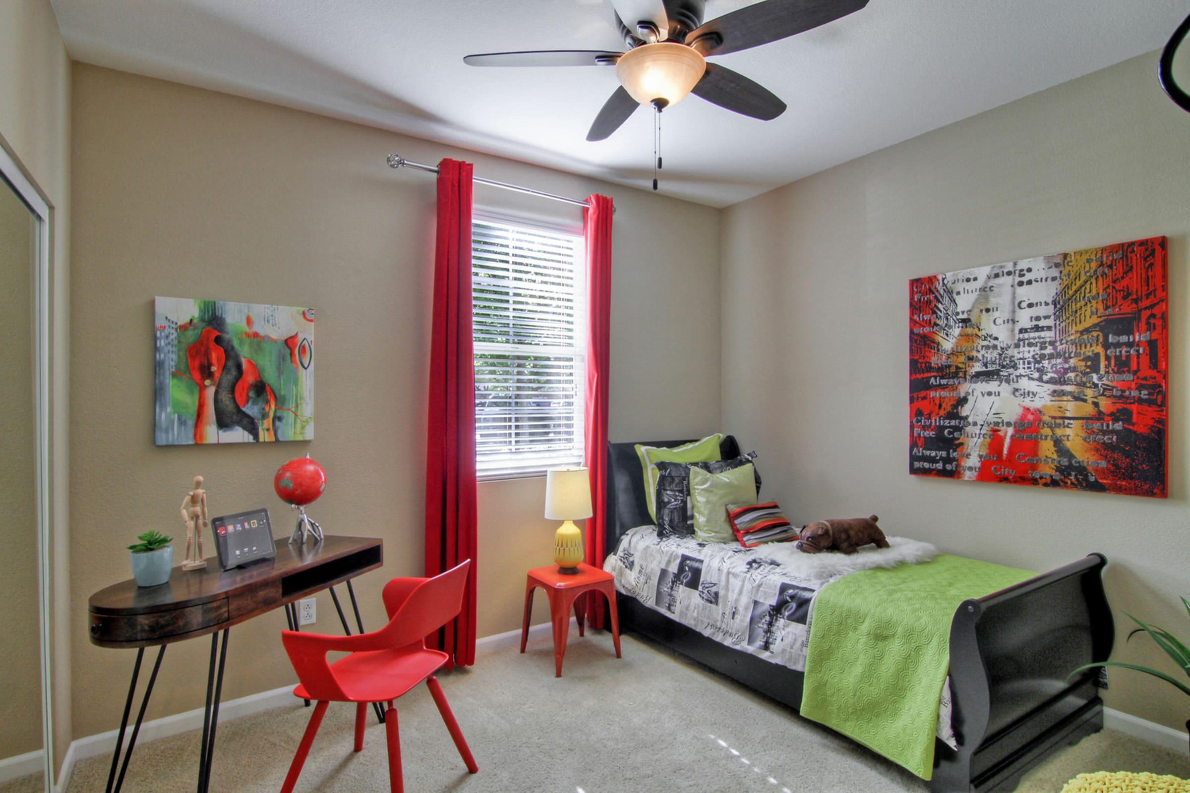A cozy bedroom featuring a single bed with colorful bedding, a small desk with a modern chair, a bedside lamp, and decorative art on the walls. Large windows with red curtains let in natural light, and a small plant adds a touch of greenery to the space.