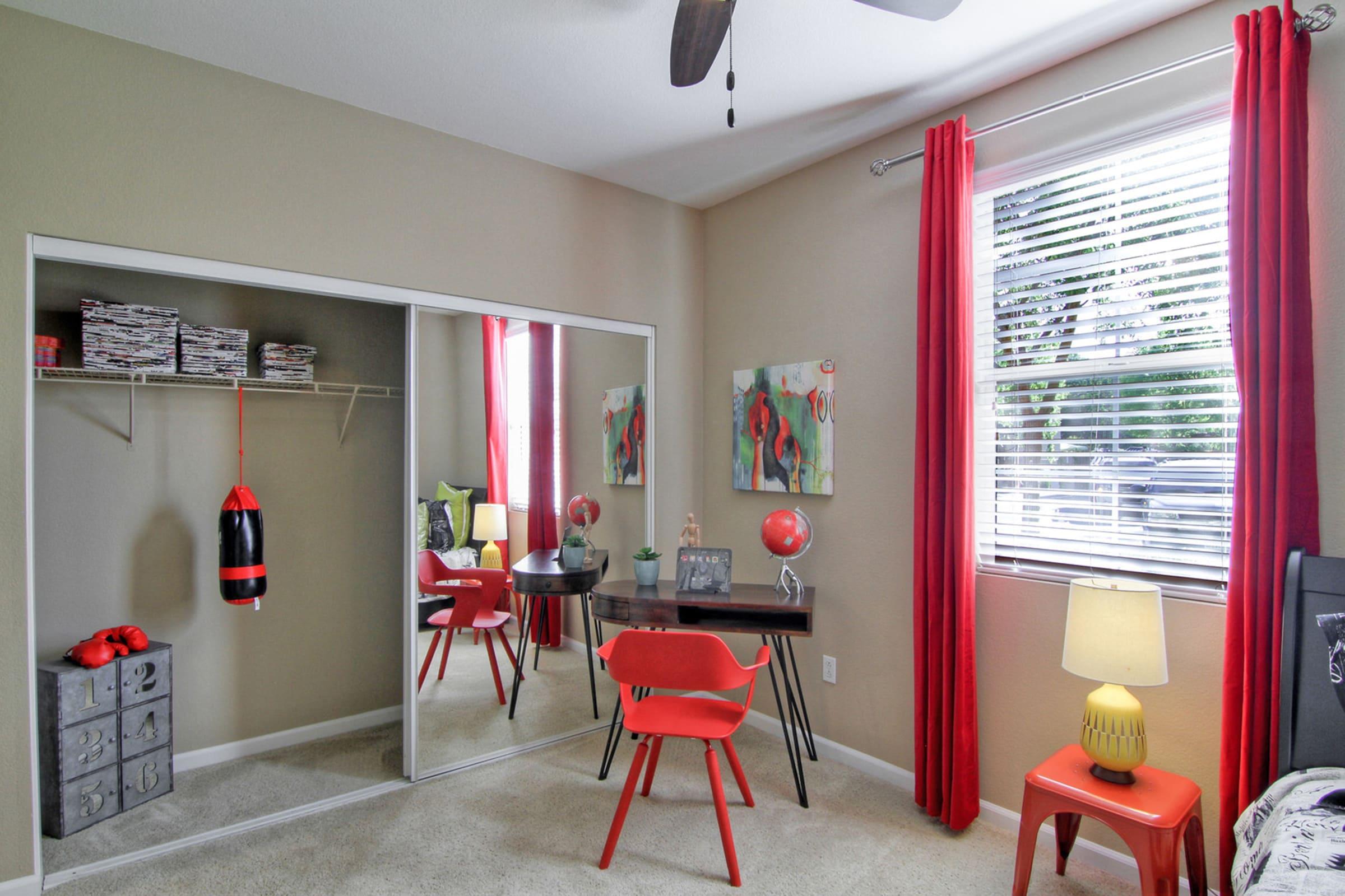 A cozy bedroom featuring a mirrored closet, a black desk with a red chair, and decorative pieces. There's a lamp on a bedside table, red curtains framing the window, and artwork on the walls. A boxing bag hangs nearby, adding a sporty touch to the room. The carpet is light-colored, creating a warm atmosphere.