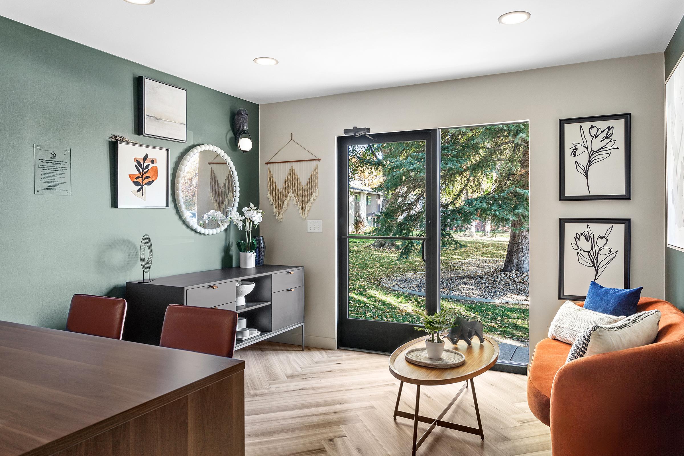 A cozy living space featuring a dark wood dining table, a stylish sideboard with decorative items, and an orange sofa. Large windows allow natural light to fill the room, showcasing an outdoor view of trees and greenery. Artistic wall decor adds character to the space.