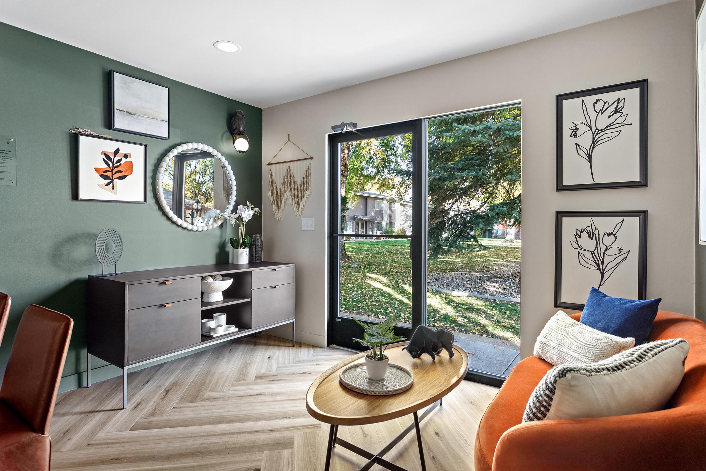 A modern living space featuring a cozy orange couch, a wooden coffee table, and a dark sideboard. The walls are painted in green and beige, adorned with framed artworks and a circular mirror. A large window offers a view of an outdoor area with trees, creating a bright, inviting atmosphere.