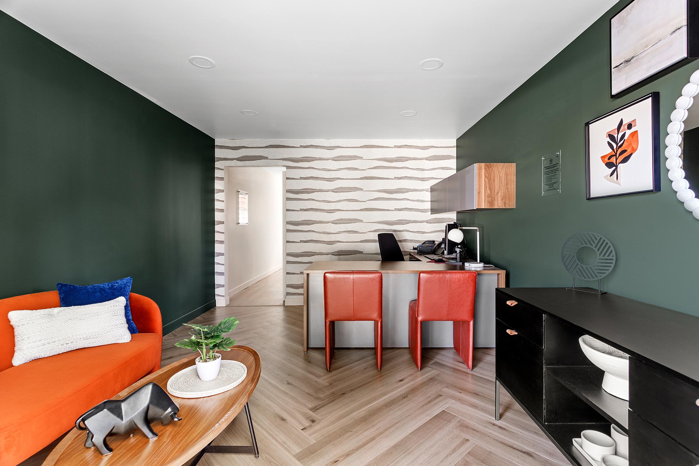 A modern office space featuring a dark green accent wall, a patterned light-colored wall behind a wooden desk with a chair, and orange leather chairs. There's a stylish orange sofa with a decorative pillow, a round mirror, framed artwork, and a coffee table with a small plant and decorative items.