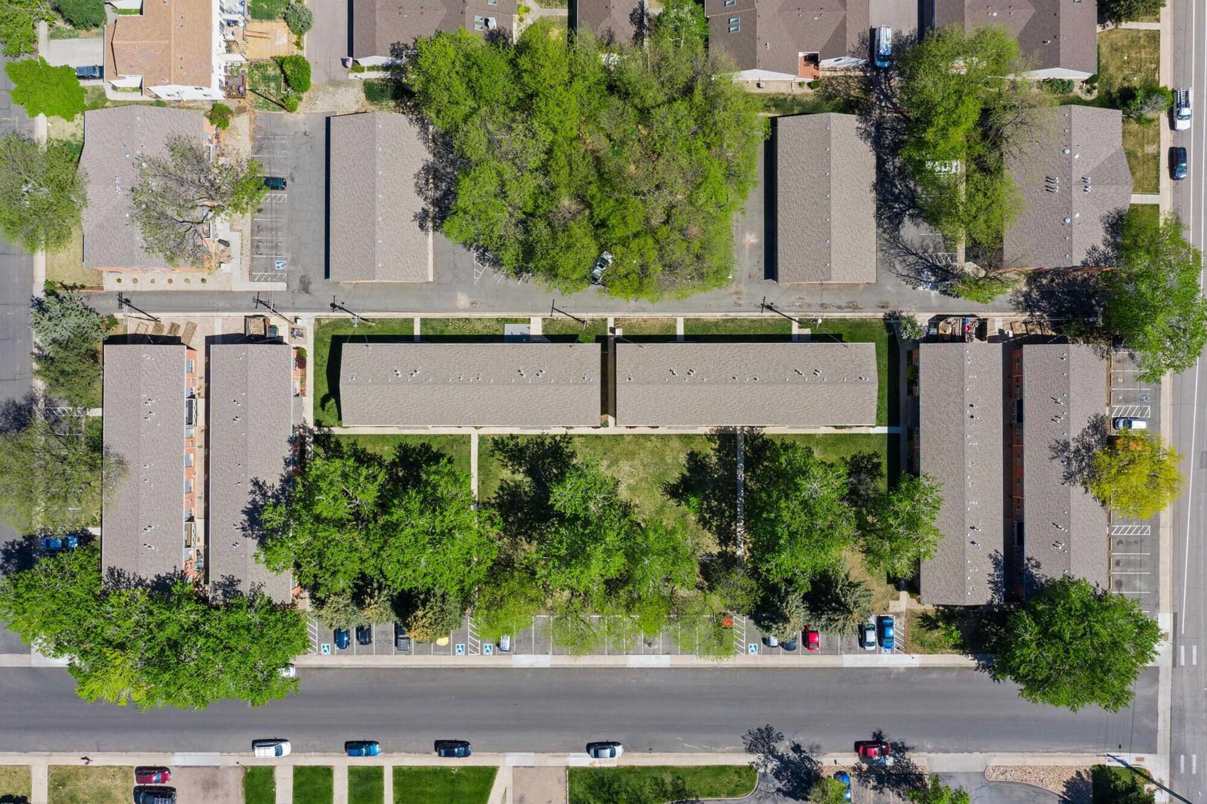 Aerial view of a residential area featuring several low-rise apartment buildings surrounded by green trees and well-maintained lawns, with a road running along the bottom and parked cars visible. The layout shows organized pathways and green spaces between the buildings.