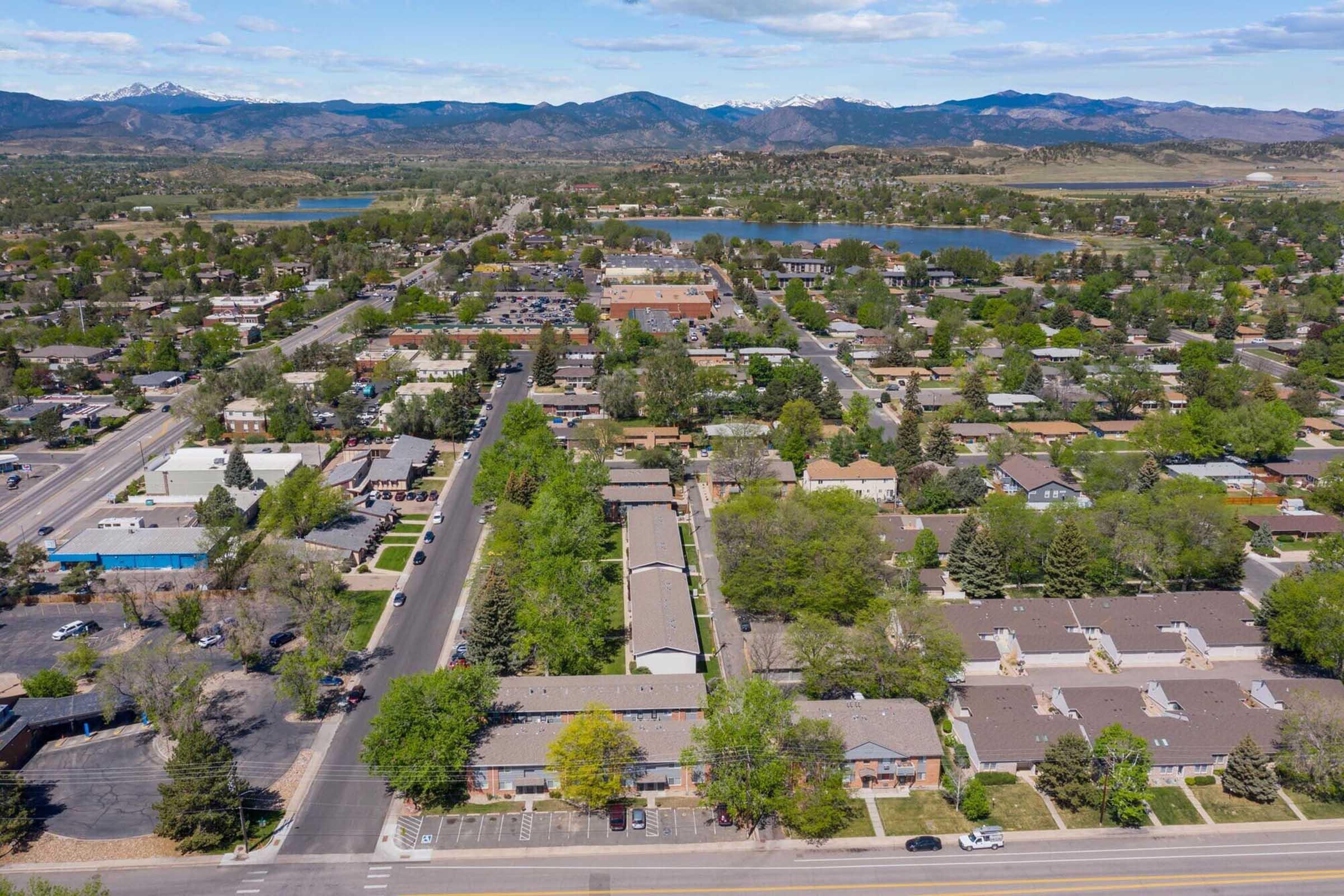Aerial view of a suburban area featuring residential neighborhoods, commercial buildings, and nearby lakes with mountainous terrain in the background. The scene is characterized by greenery and open spaces, showcasing a blend of urban development and natural landscapes.