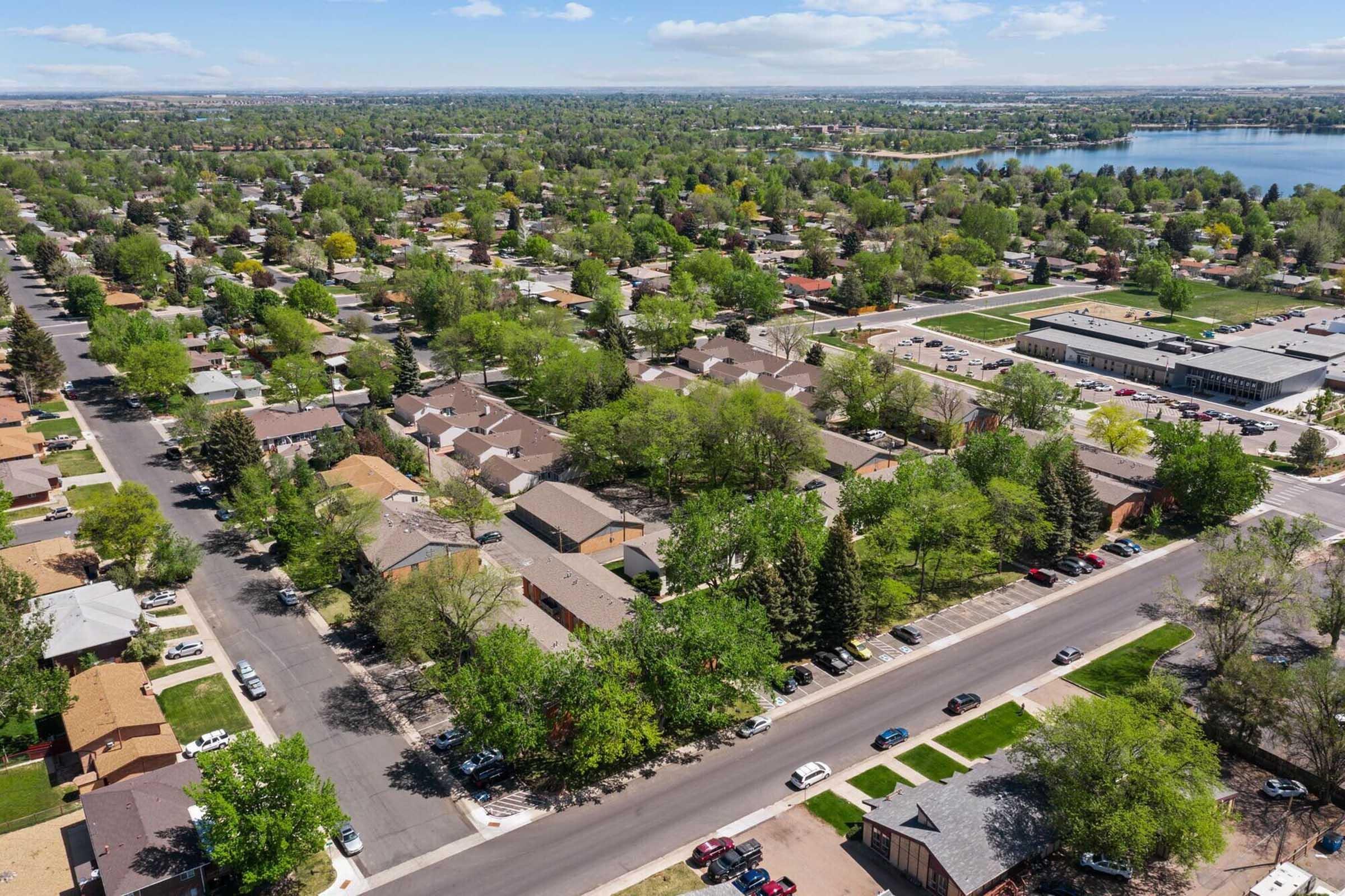 Aerial view of a suburban neighborhood featuring well-maintained homes, tree-lined streets, and green spaces. A large lake is visible in the background, surrounded by trees, and several cars parked along the streets. The scene captures a picturesque, residential area with a blend of greenery and urban elements.