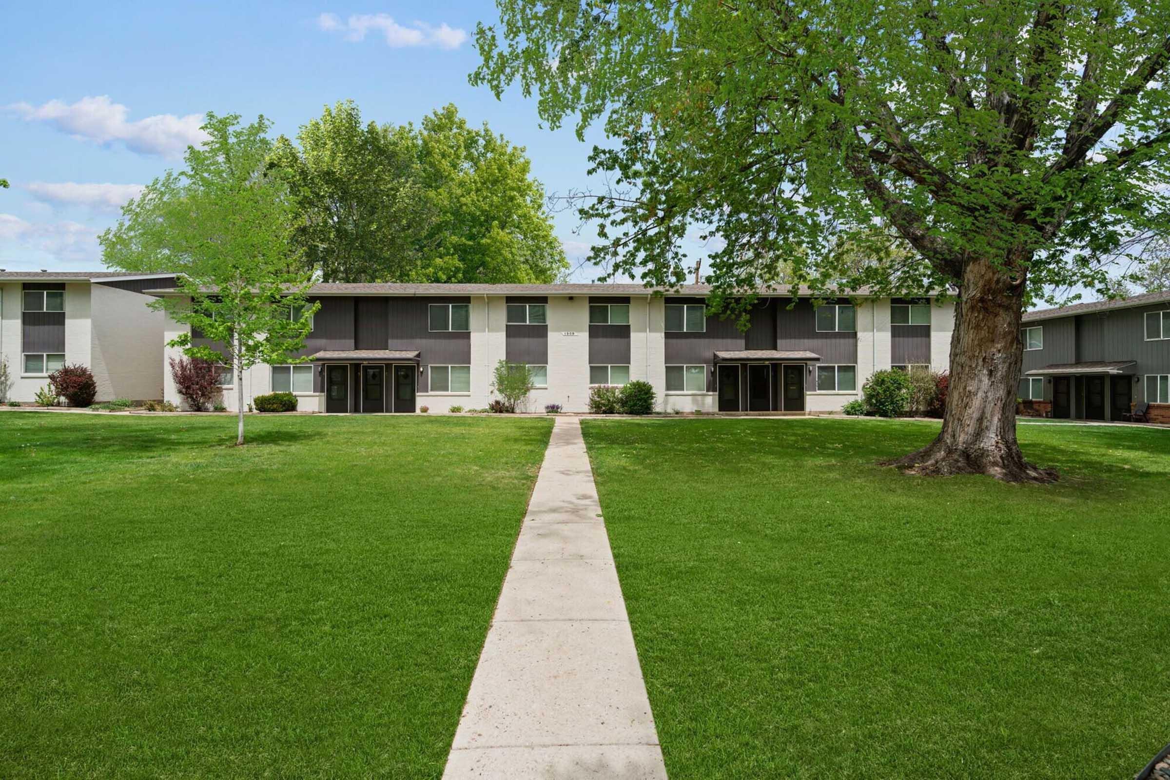 A well-maintained residential complex featuring multiple buildings with modern architecture, surrounded by lush green lawns and trees. A clean, paved pathway leads through the grassy area, inviting residents to stroll. The sky is clear with a few clouds, enhancing the peaceful atmosphere of the neighborhood.