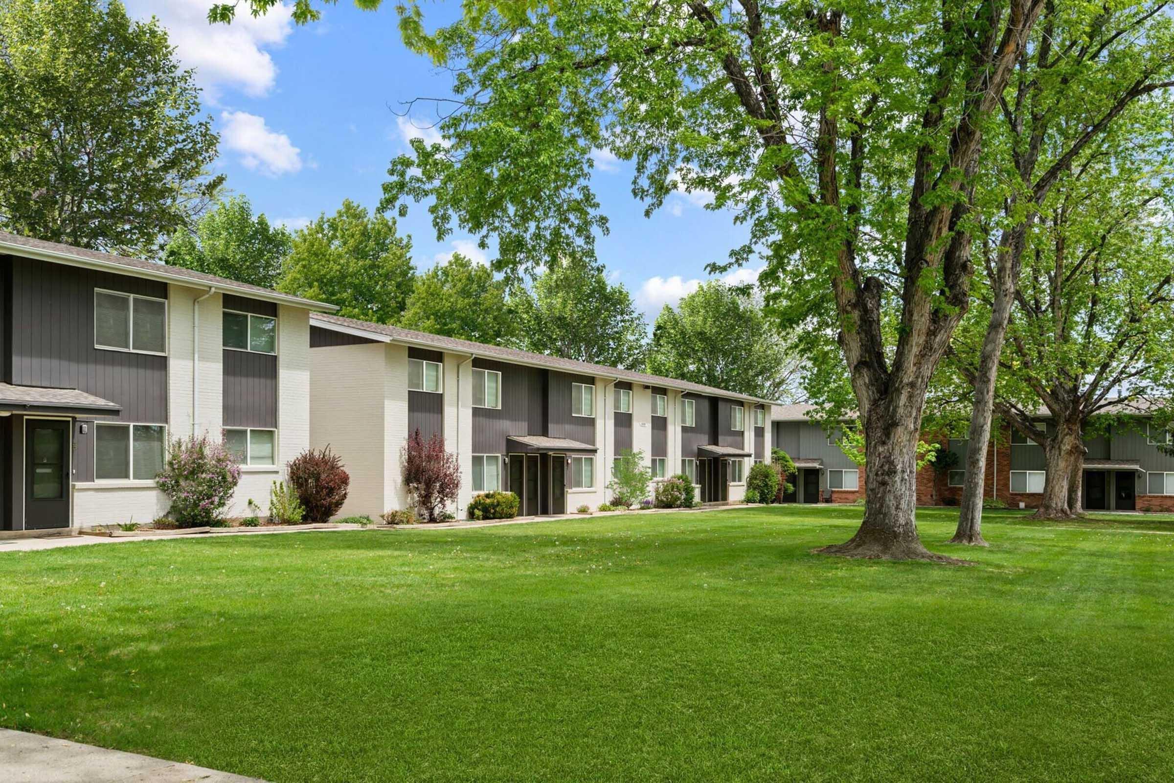 Row of apartment buildings surrounded by lush green grass and trees under a blue sky with scattered clouds. The buildings feature a mix of white and dark siding, with landscaped shrubs in front. Ideal for a peaceful residential setting.